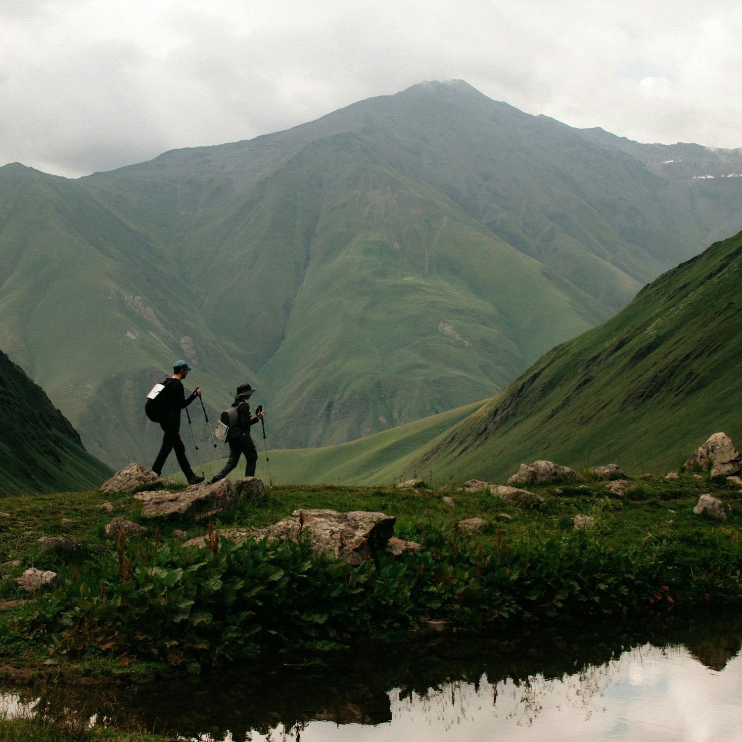 A man and woman hike with trekking poles by a mountain lake.