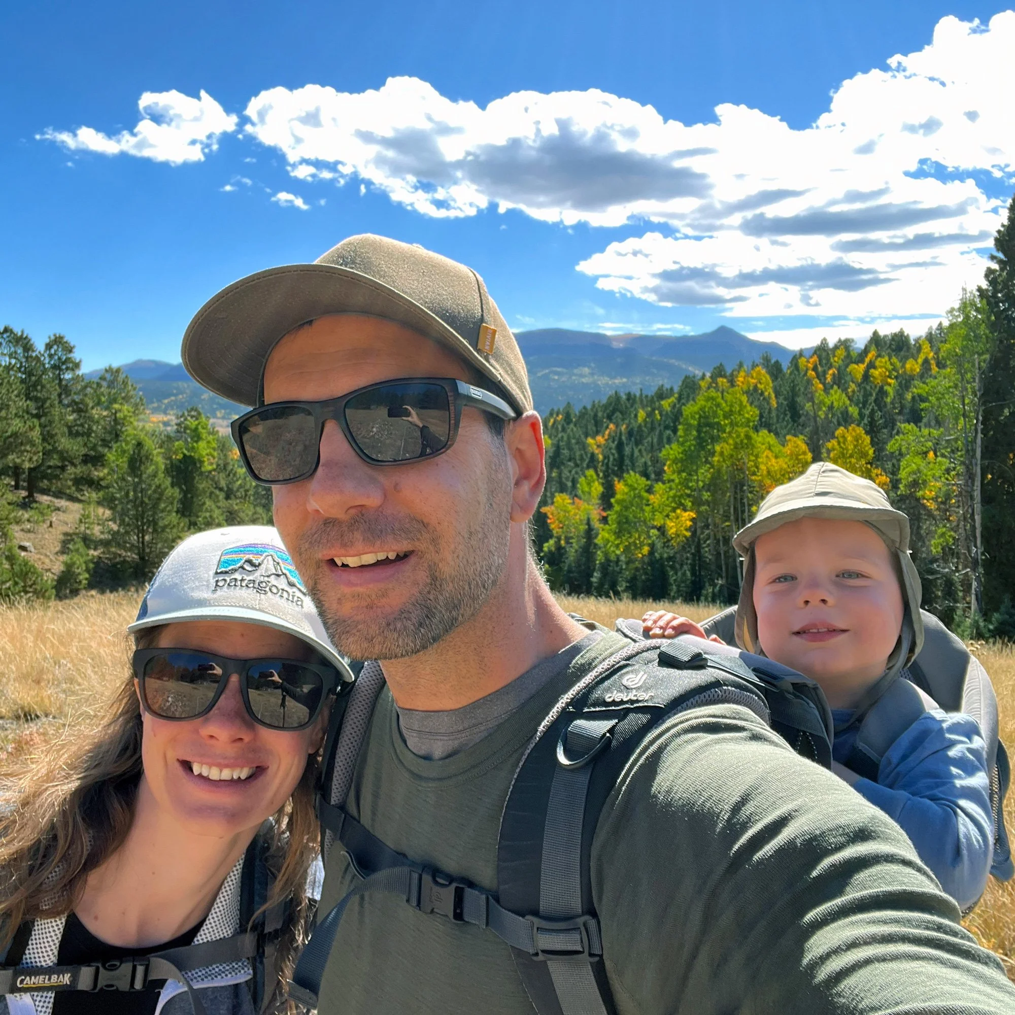 A family of three enjoying a hike outdoors with mountains and a blue sky with clouds in the background. The man is wearing sunglasses and a cap, the woman is wearing sunglasses and a hat, and the child is in a backpack carrier.