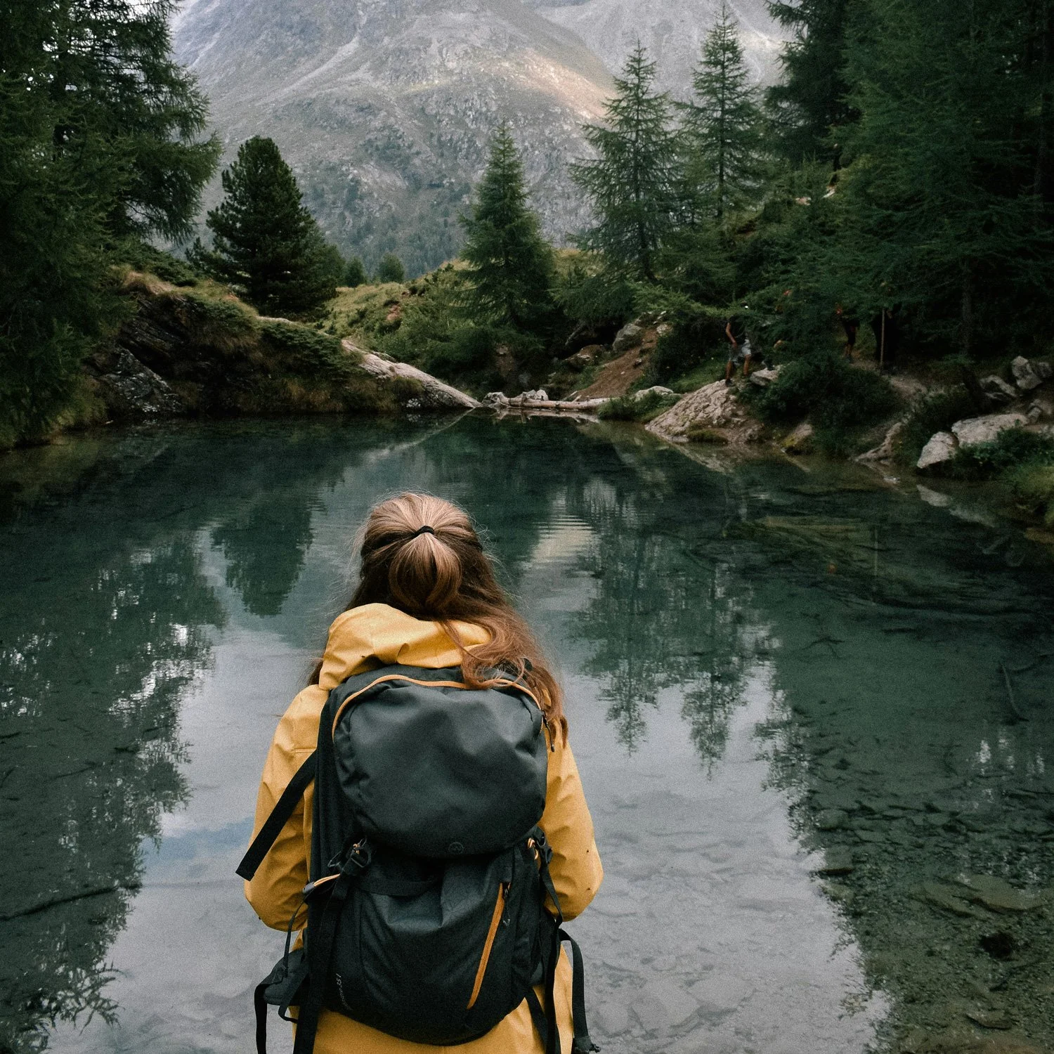 Girl stands in front of a mountain lake with a small backpack.