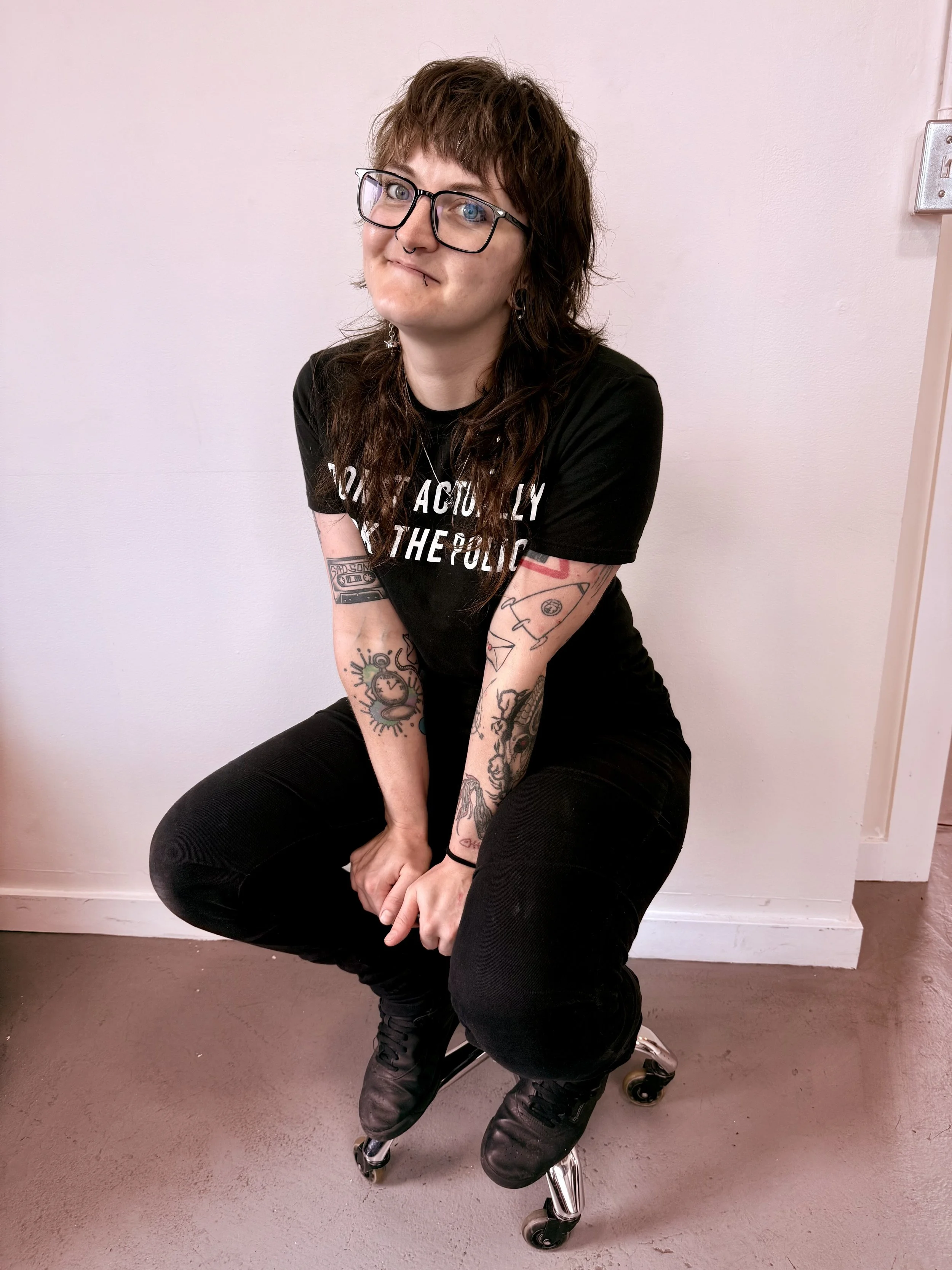 A person with brown shaggy hair, glasses, and tattoos on their arms, sitting on a caster stool against a plain white wall. They are wearing a black t-shirt, black pants, and black shoes, and is smiling at the camera.