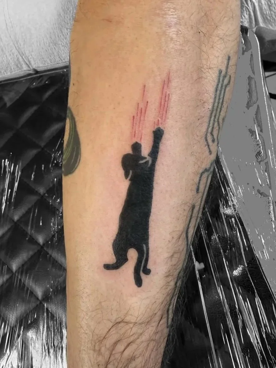 Black silhouette of a cat caught mid-air, with red lines indicating motion or impact, on a person's forearm.