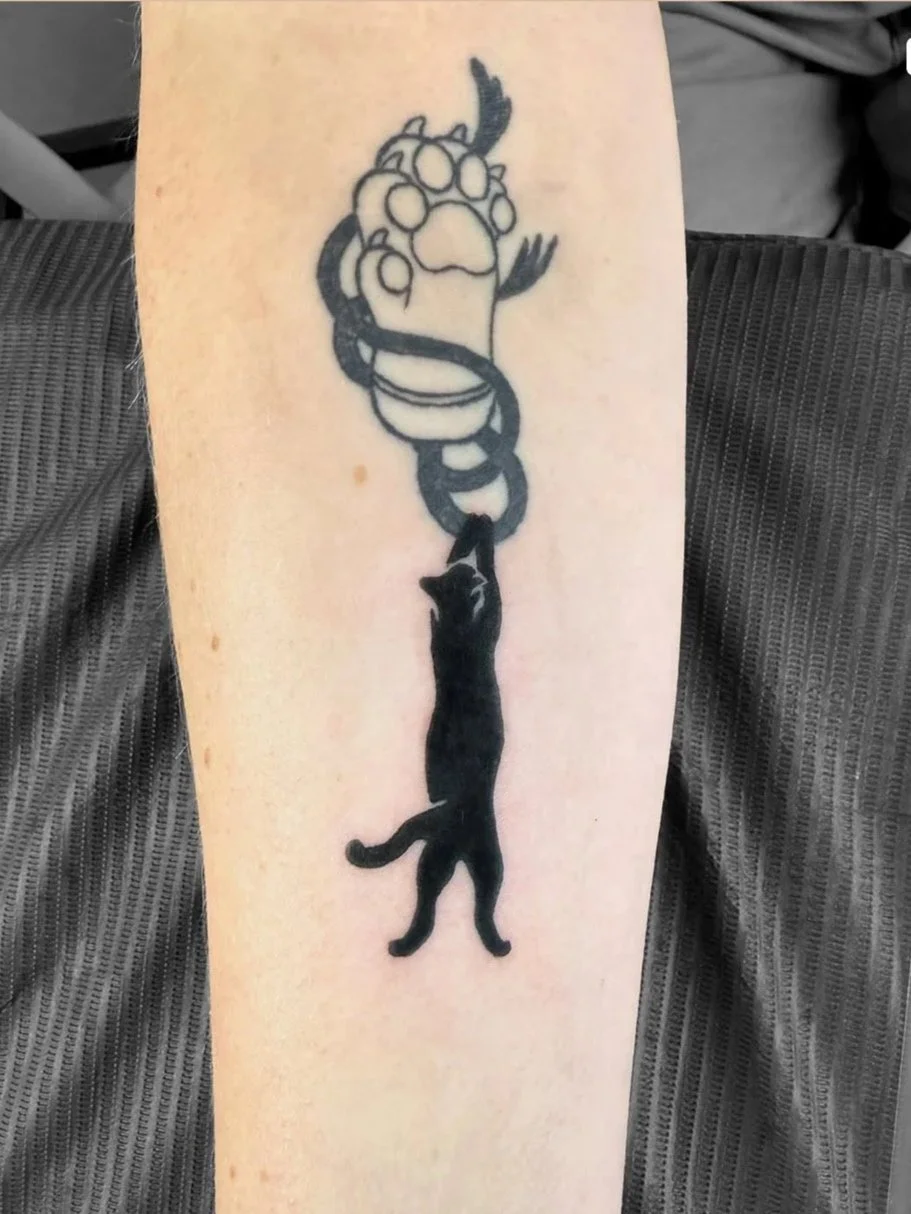 A tattoo of a black cat hanging from a paw print, with a snake intertwined around its body.