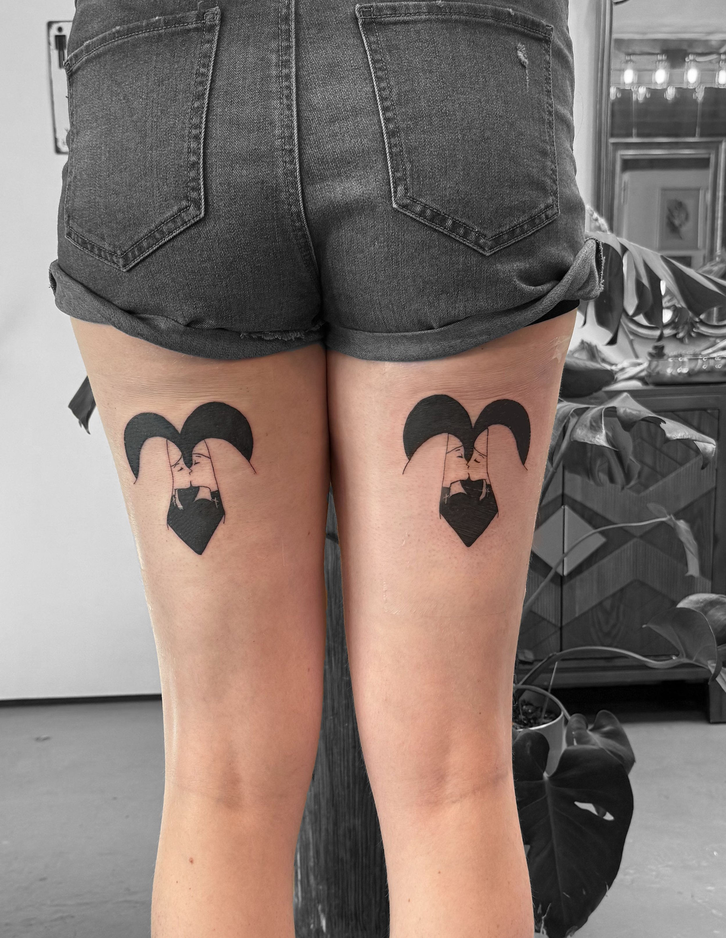 Tattoo of two women kissing, with one woman having long hair and the other with a swimsuit. The tattoo is black and located on the person's upper thighs.