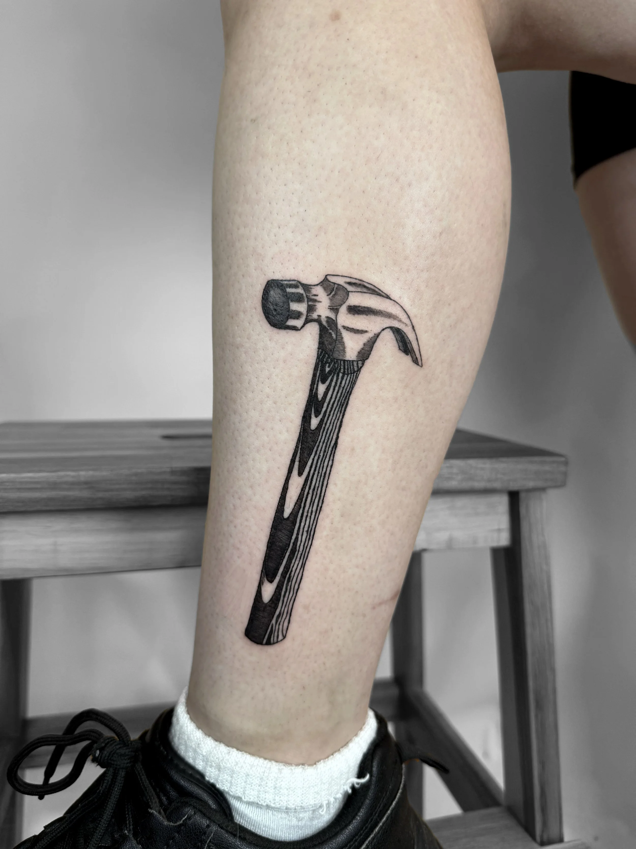 Tattoo of a hammer with a wooden handle and metal head on a person's leg.