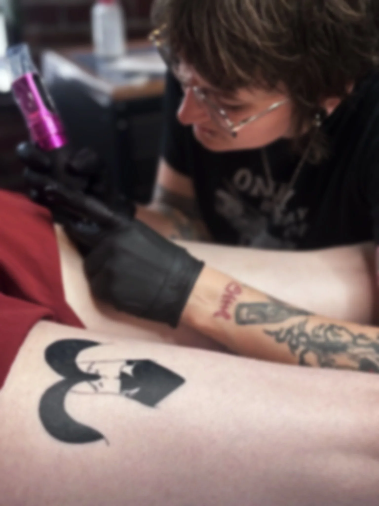 Person with glasses and tattoos is tattooing a design on someone else's arm in a tattoo shop.