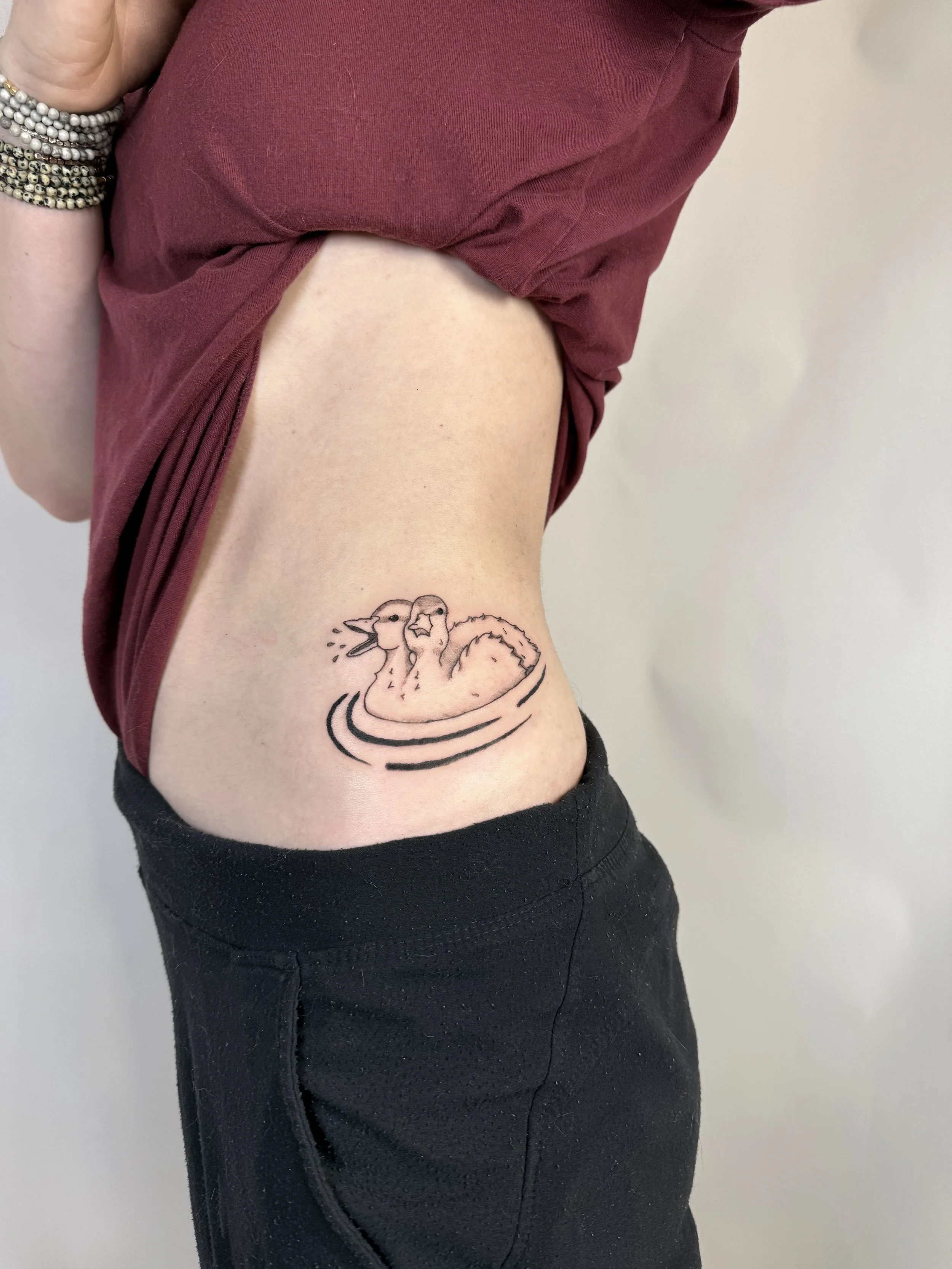Tattoo of two ducks swimming in water on person's side ribcage.