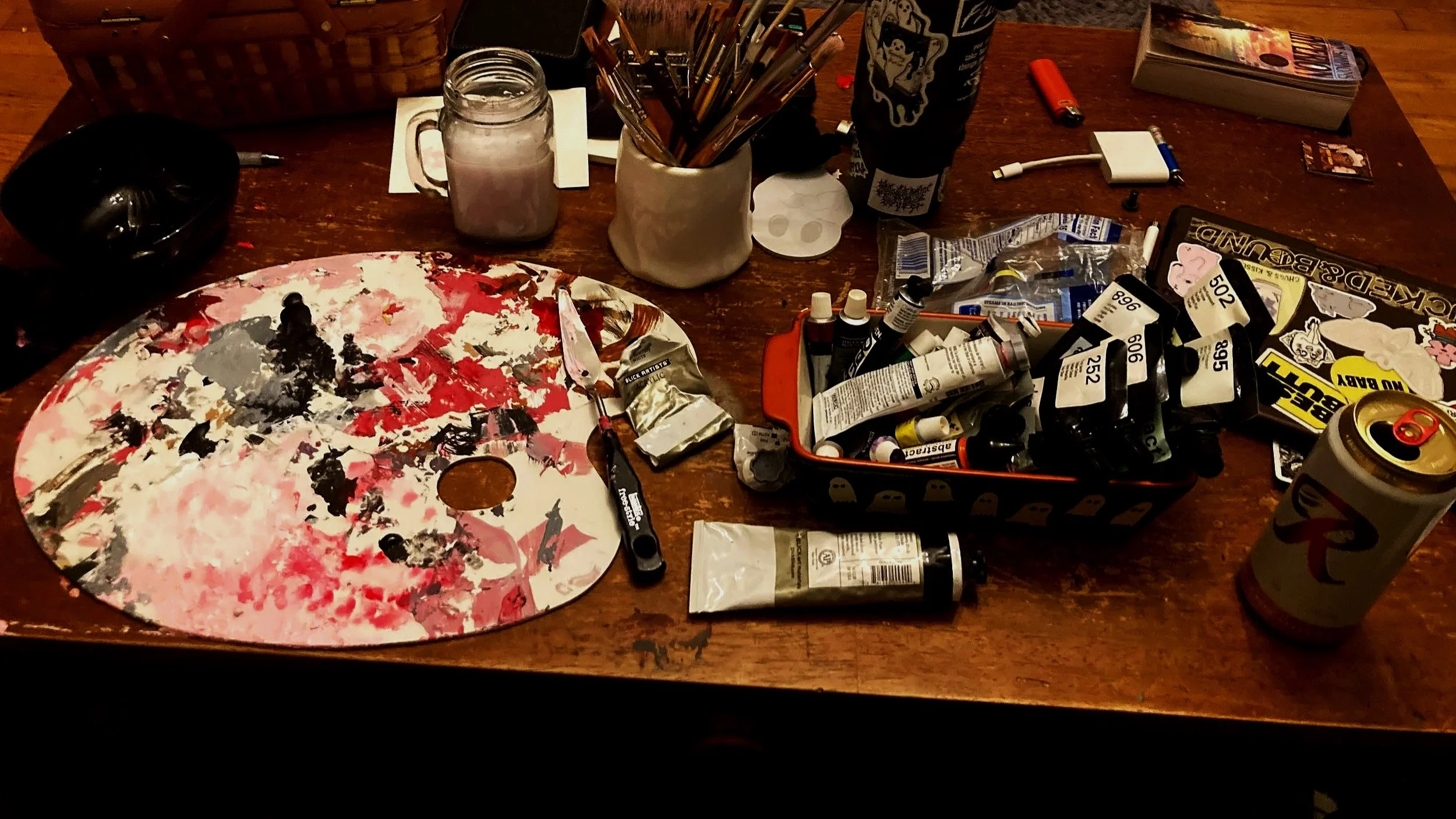 An art workspace with a colorful paint palette, tubes of paint, brushes in a white cup, a black bowl, a small cutting knife, a tube of paint, a lottery ticket, a soda can, a book, and various art supplies scattered on a wooden table.