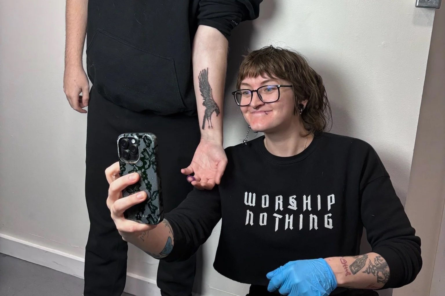 A person with glasses and tattoos on their arms taking a selfie, while a person standing next to them shows their tattooed wrist.