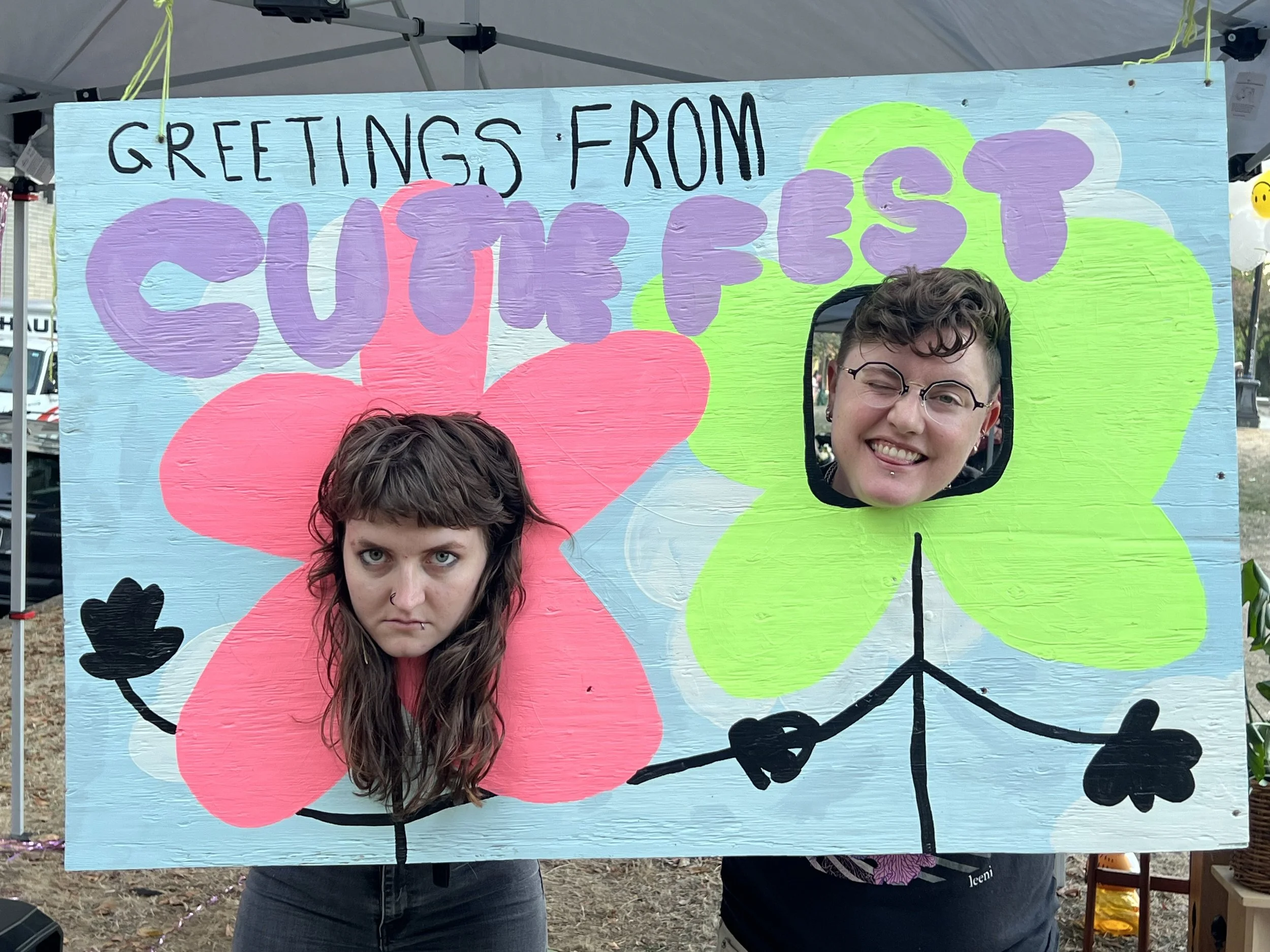 A person with long brown hair and a serious expression is sticking their head through a cutout in a colorful wooden board painted to look like a pink flower, while another person with short curly hair, glasses, and a big smile is using the cutout of a green flower to make a funny face. The board has the text 'Greetings from Sunwest' written on it and is decorated with flowers and hearts.