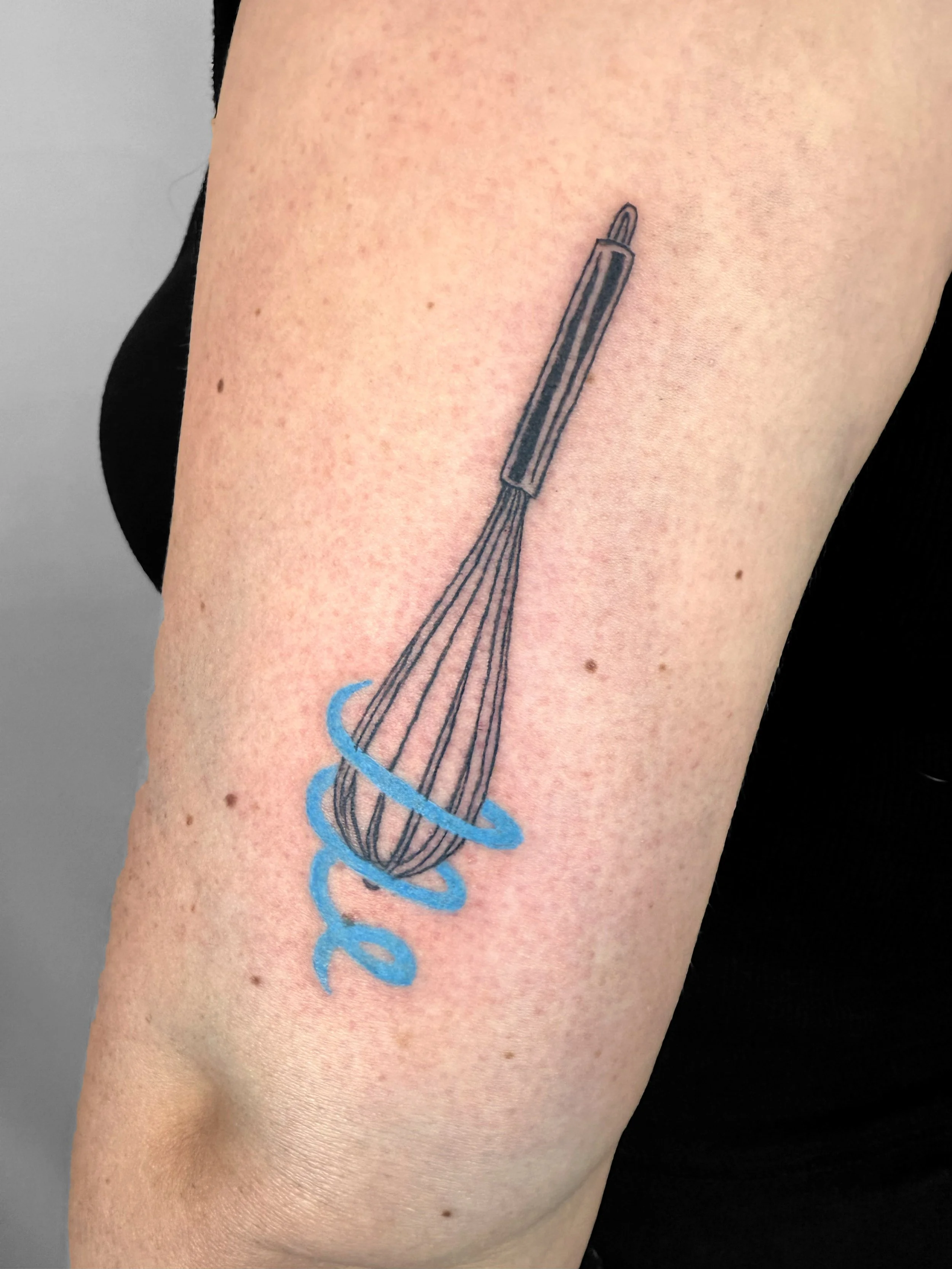 Tattoo of a whisk with the word 'dope' written in blue below it on person's upper arm.