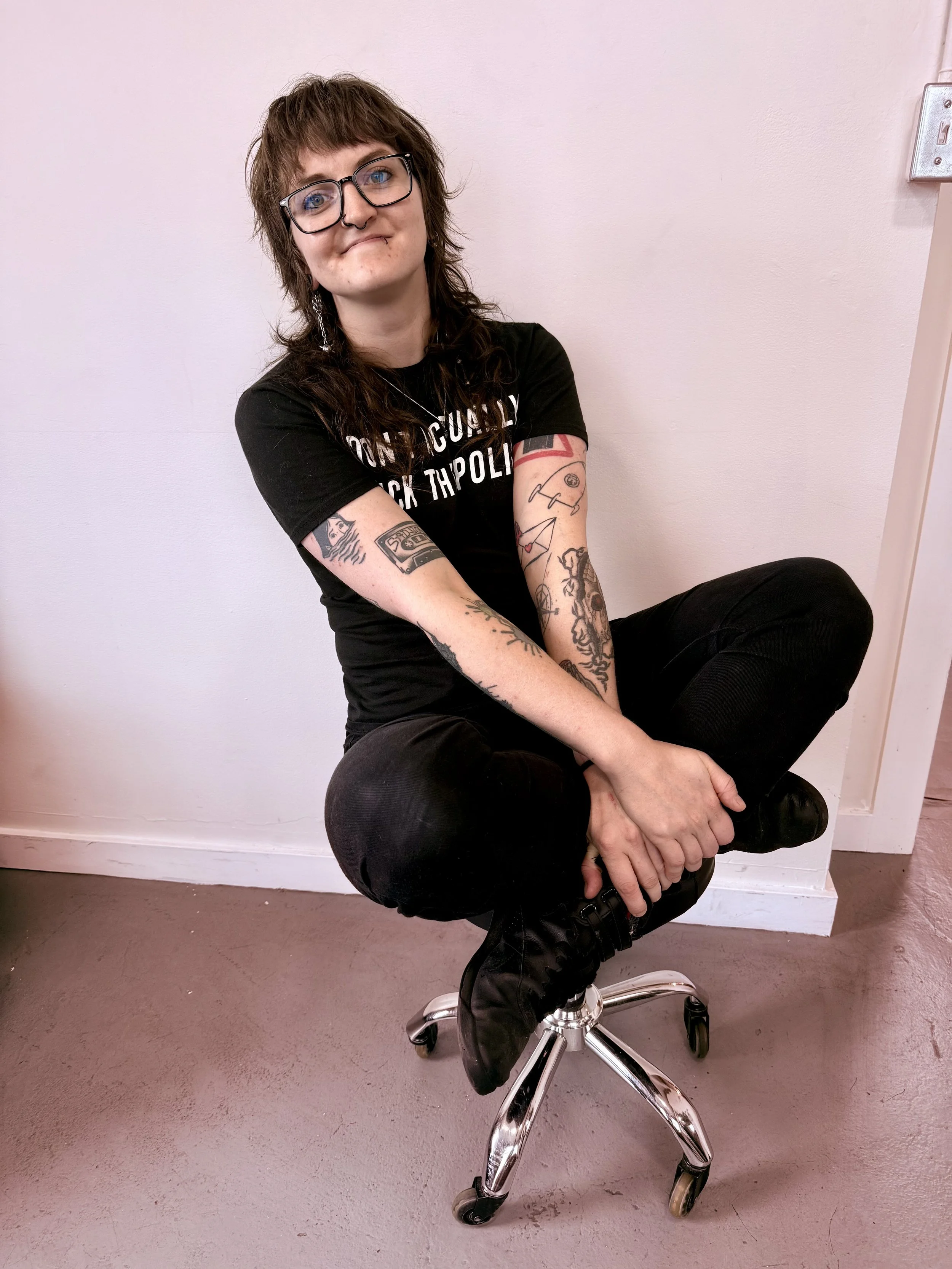 A person with glasses and brown, shoulder-length hair sitting cross-legged on a swivel stool, wearing a black T-shirt with white text, black jeans, and black boots, with visible tattoos on both arms, against a white wall.