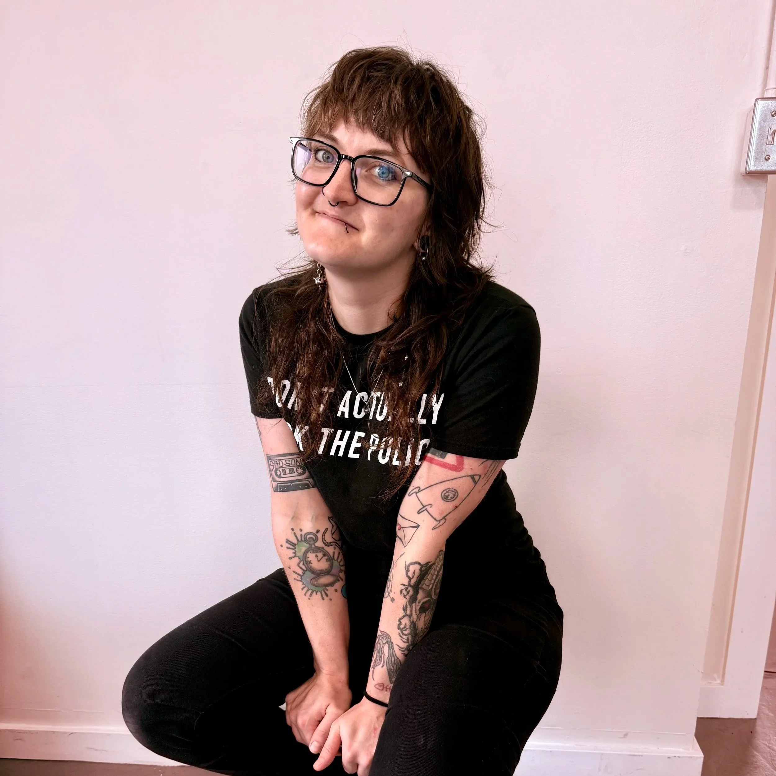 Person with brown hair, glasses, and tattoos, sitting against a plain wall, wearing a black t-shirt and black pants, smiling slightly.
