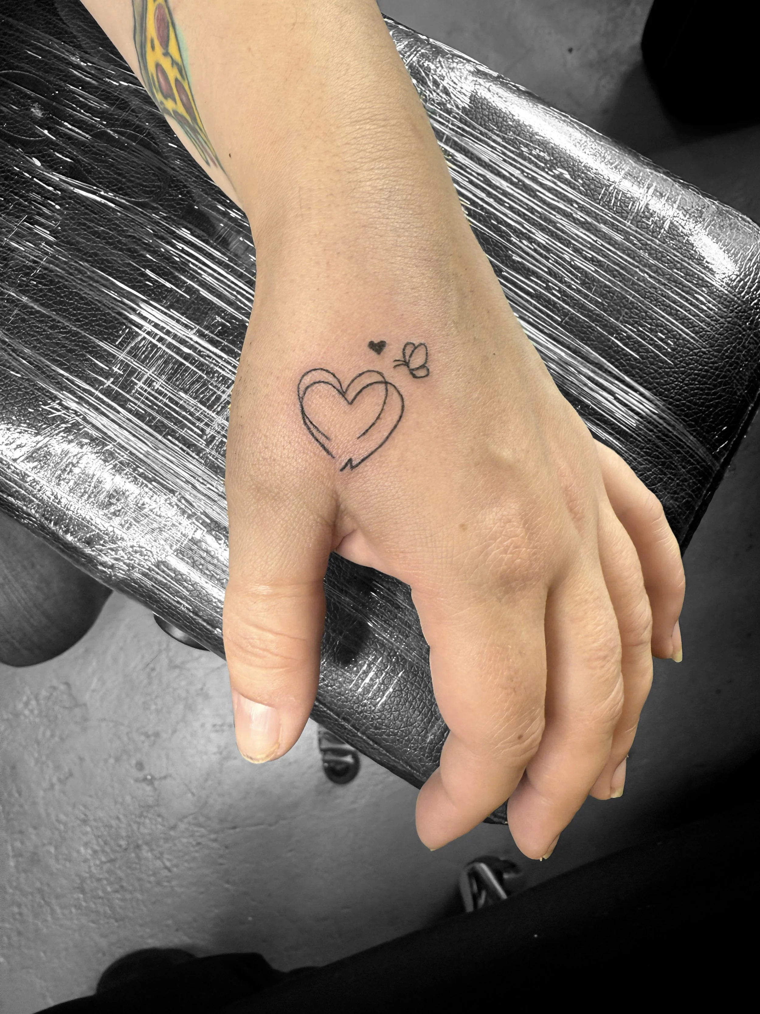 A hand with a tattoo of a broken heart, a small solid black heart, and a butterfly on the wrist, resting on a black leather surface.