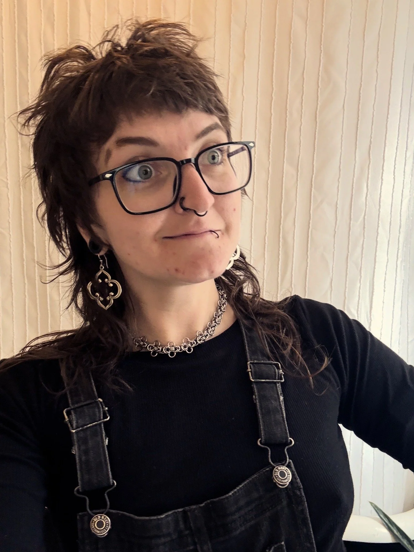 Person with short spiky hair, glasses, and multiple piercings, wearing a black shirt and overalls, posing indoors against a textured cream-colored wall.