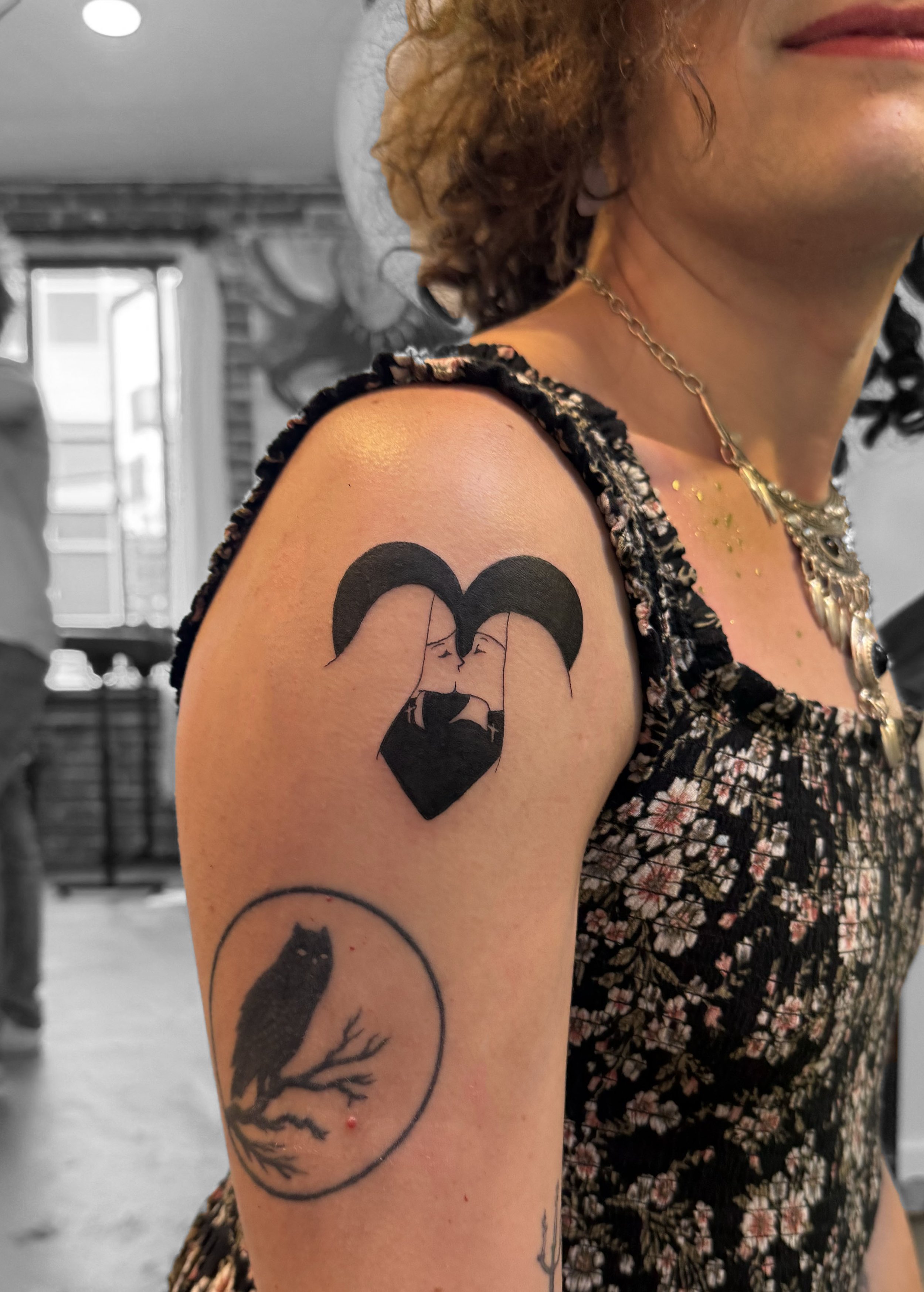 Tattoo of two women with long dark hair kissing, forming a heart shape, on a person's upper arm. Below it, a tattoo of a black owl sitting on a branch inside a circle.