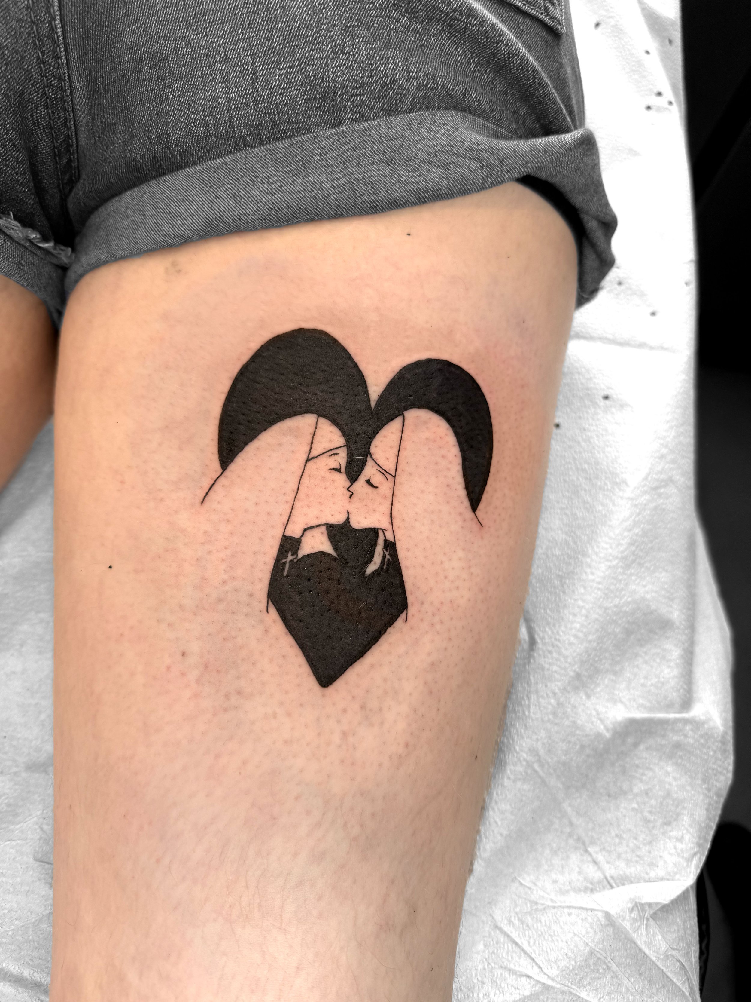 Tattoo of two women with black hair, facing each other, forming a heart shape with their heads and shoulders, with a black heart shape below them