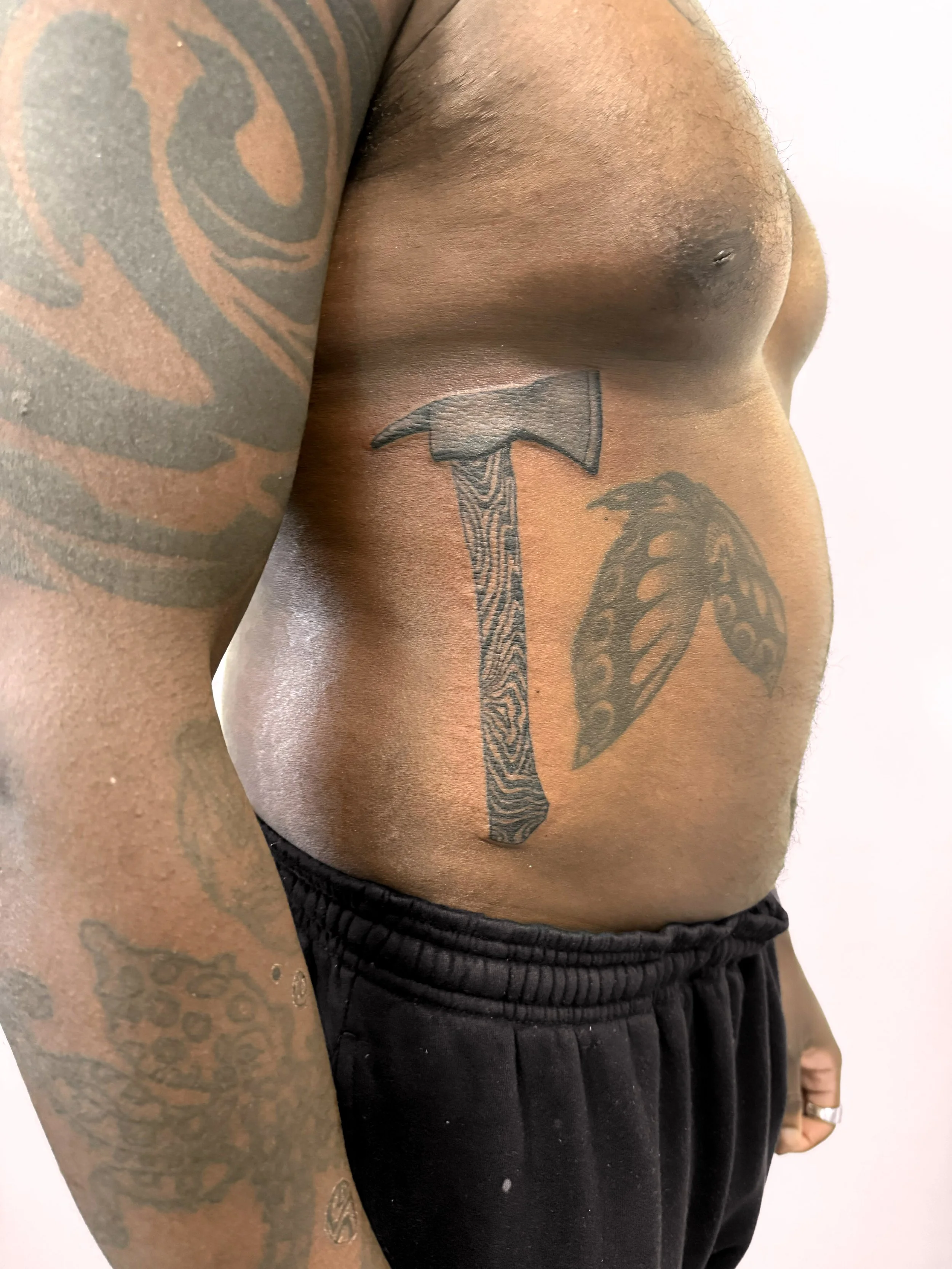 Close-up of a person's chest and side, showing tattoos of an axe and a butterfly wing. The person is shirtless and wearing black shorts.