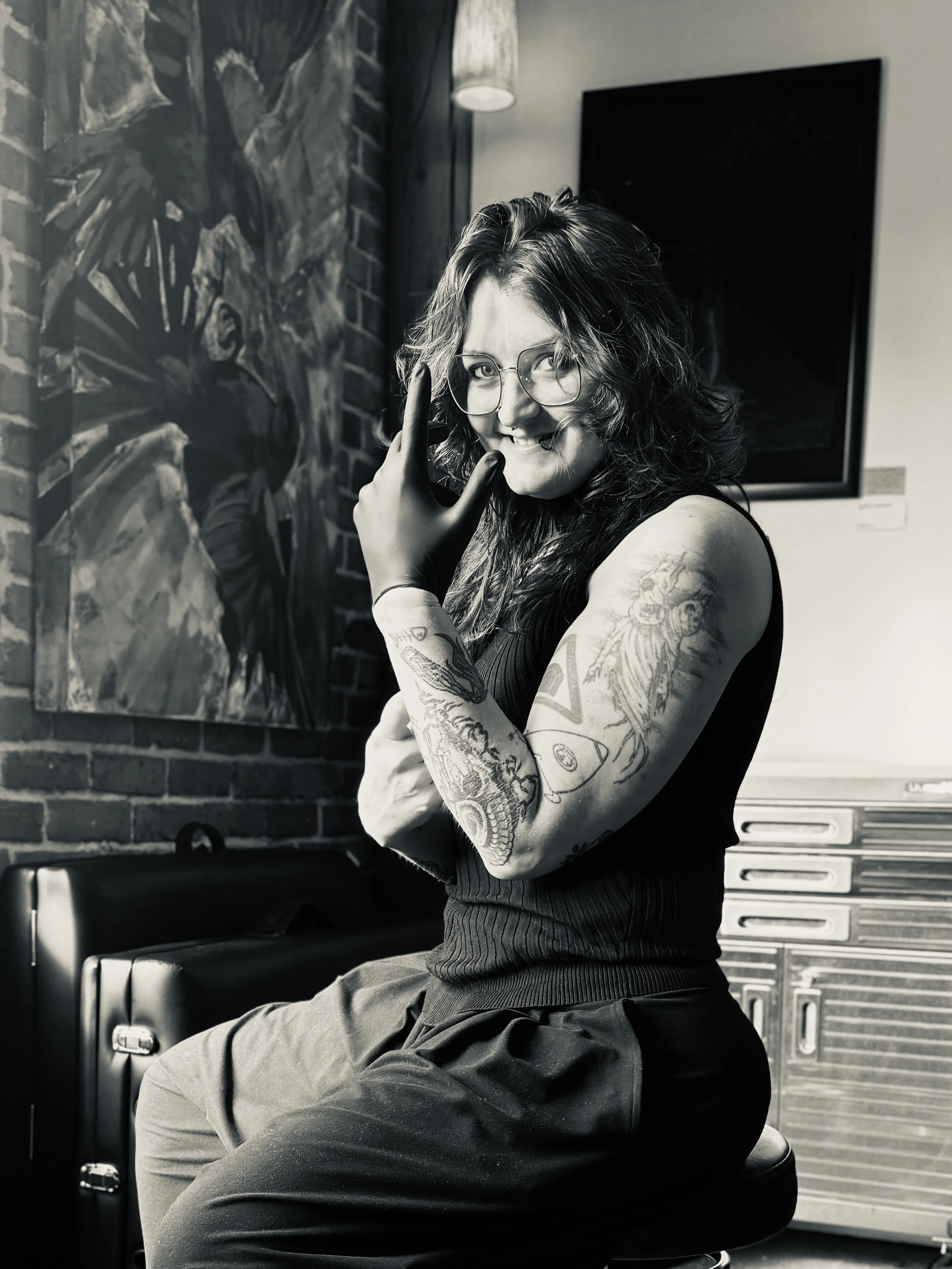 A person with curly hair and tattoos on their arms sitting on a stool, making a rock and roll hand gesture, smiling, in a modern room with artwork on the wall.