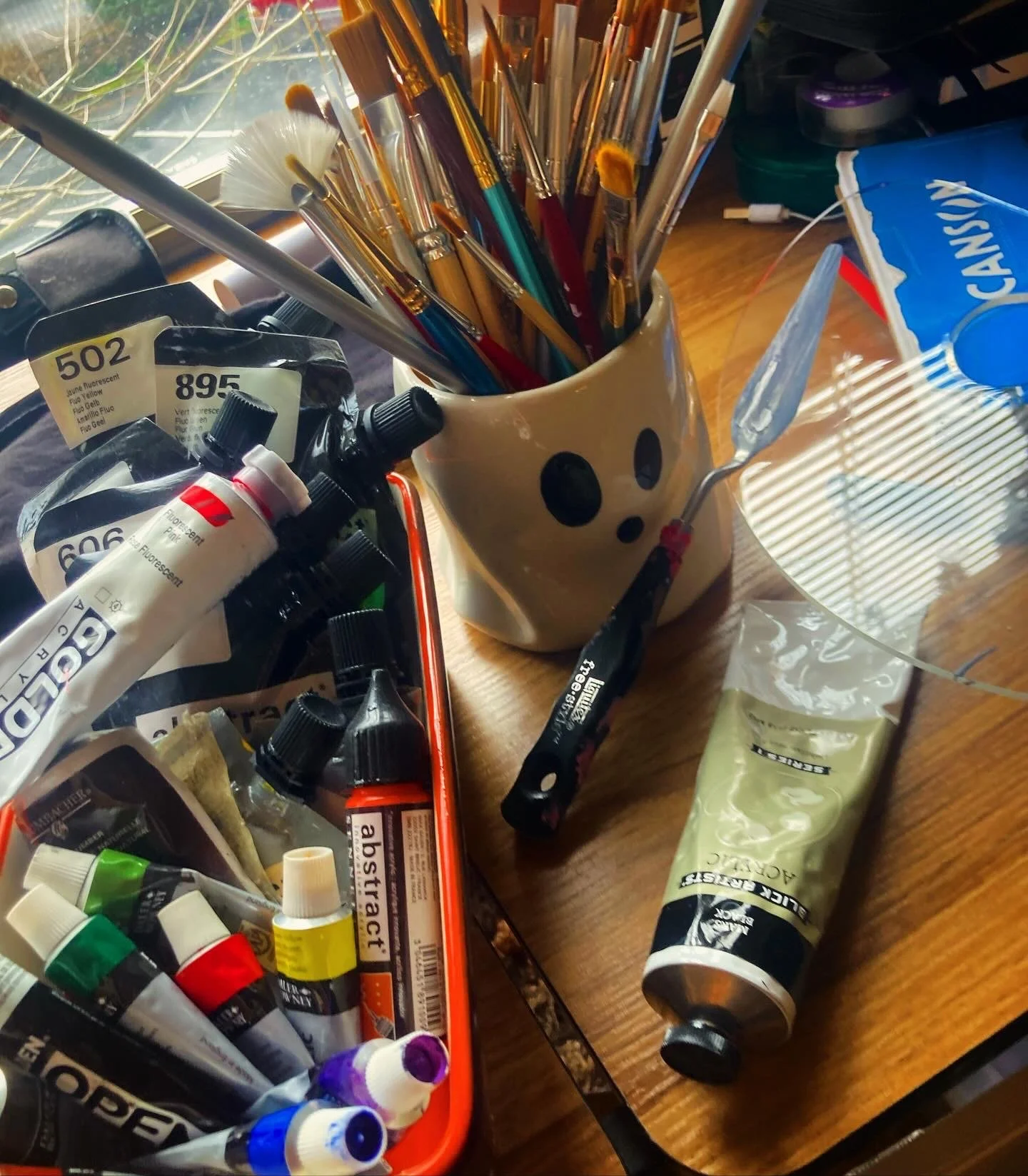 A collection of art supplies including paint tubes, brushes in a panda mug, a palette knife, and a paint mixing tray on a wooden table.
