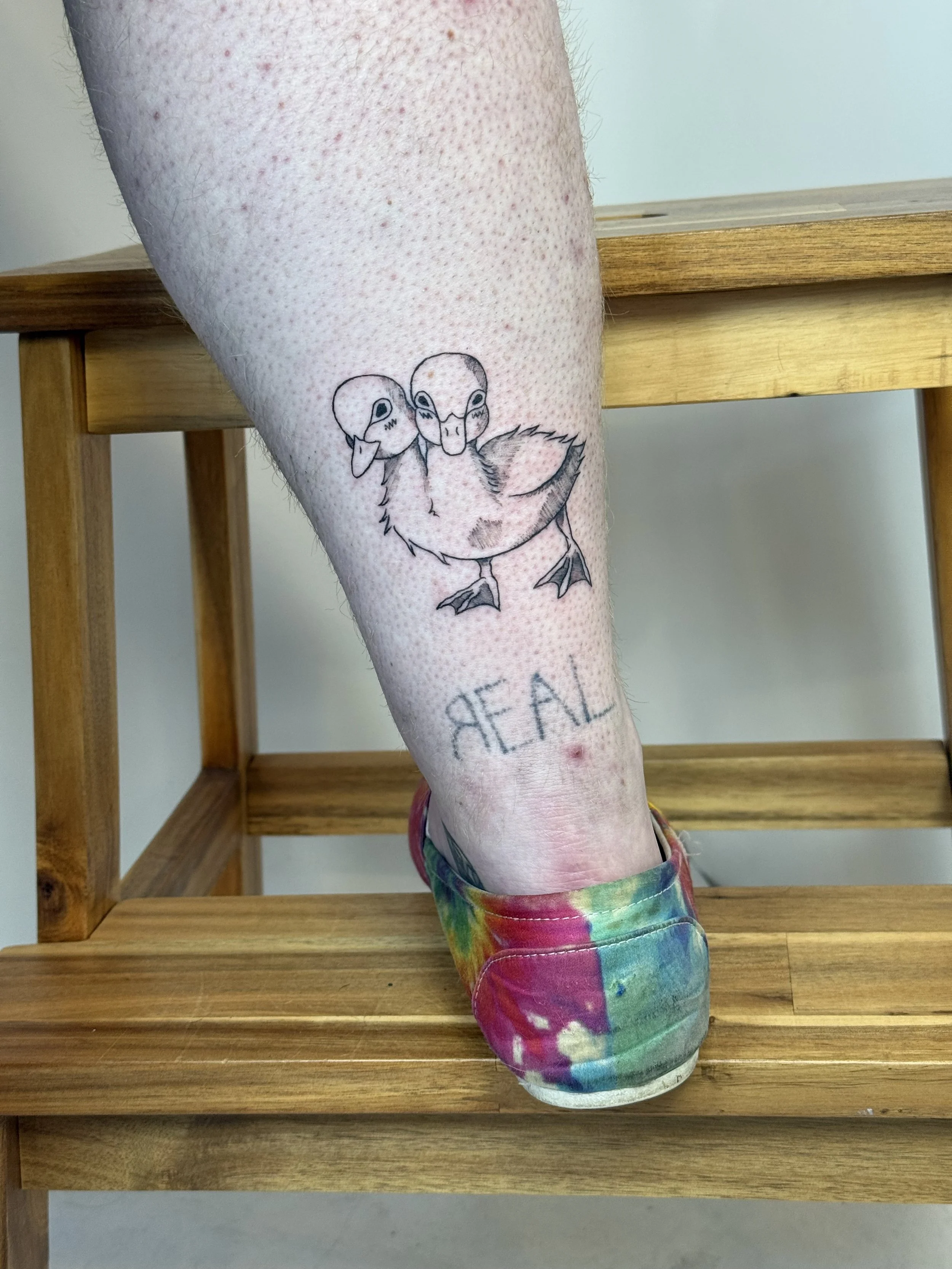 Tattoo of two ducklings with the word "REAL" underneath on a person's lower leg, standing on a wooden stool with a colorful tie-dye shoe.
