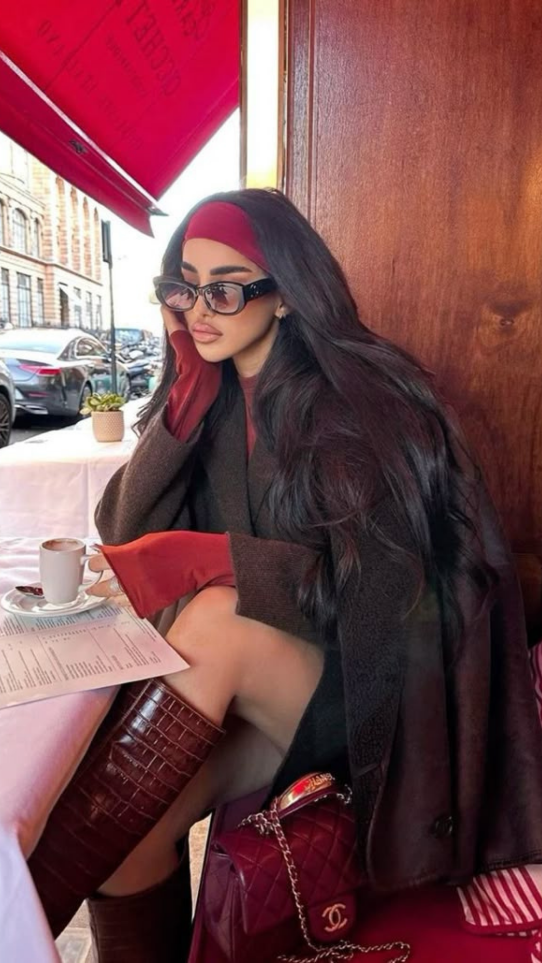 A woman sitting at an outdoor cafe table, wearing sunglasses, a headband, and a dark outfit, with long dark hair, holding a phone near her ear, and a cup of coffee on the table.