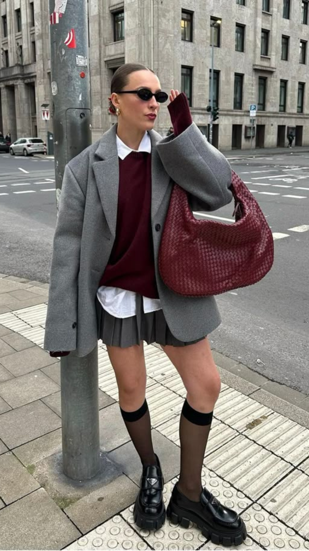 Fashion-forward woman standing on city sidewalk with oversized gray blazer, layered burgundy sweater, white shirt, pleated mini skirt, sheer black knee-high socks, chunky black loafers, sunglasses, gold earrings, and carrying a large red bag.