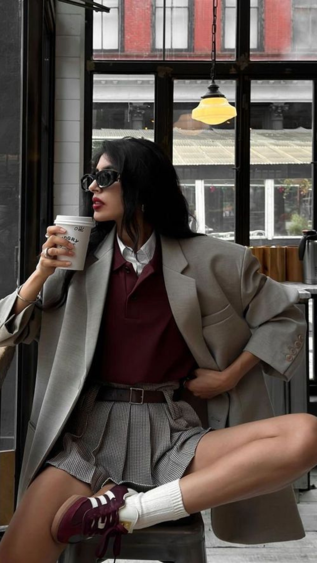 Woman wearing sunglasses, a beige blazer, maroon polo shirt, plaid shorts, sitting in a cafe, holding a coffee cup, with a window in the background.