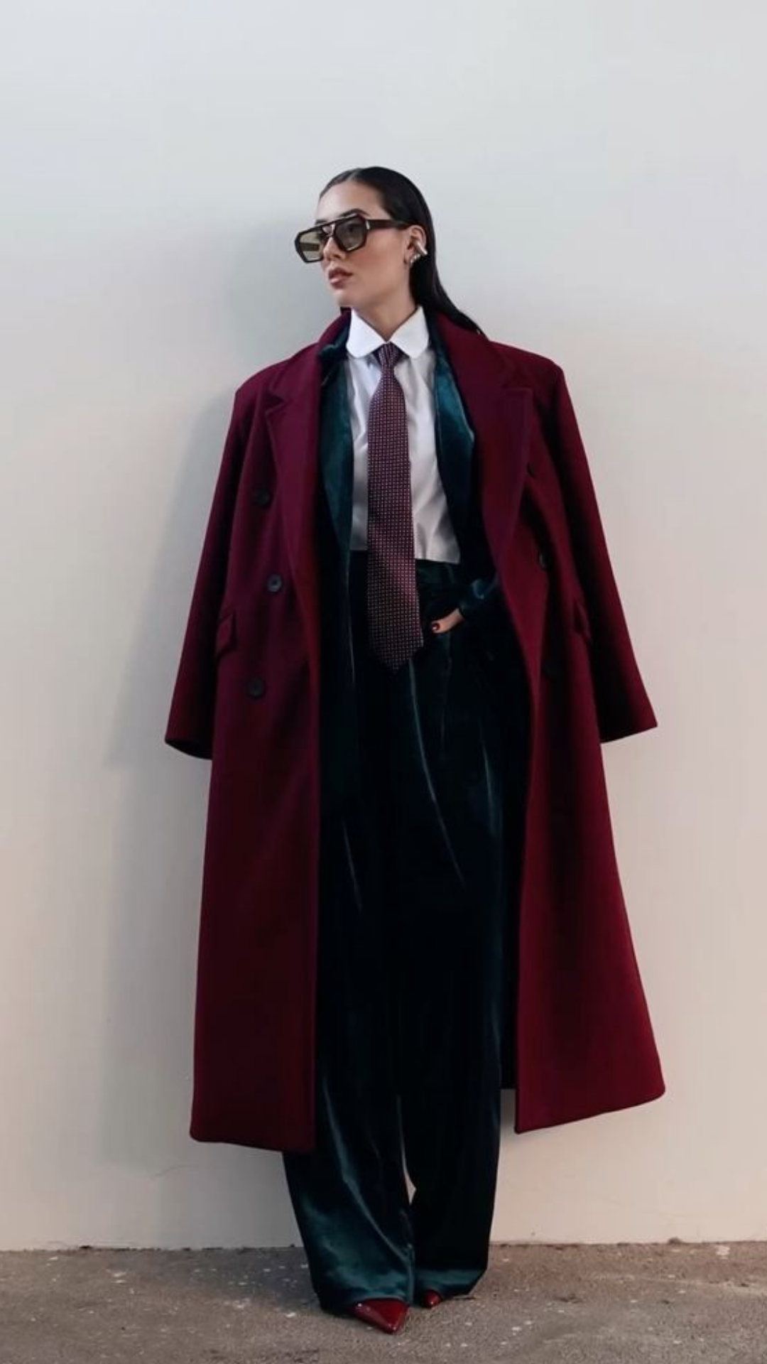 Woman wearing a long red coat, black pants, white shirt, patterned tie, black sunglasses, and red shoes, standing against a plain light-colored wall.