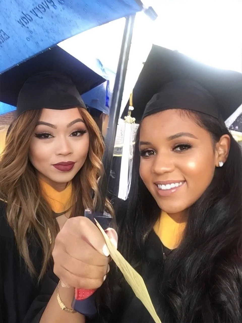 Two young women in graduation caps and gowns taking a selfie, one holding a diploma.