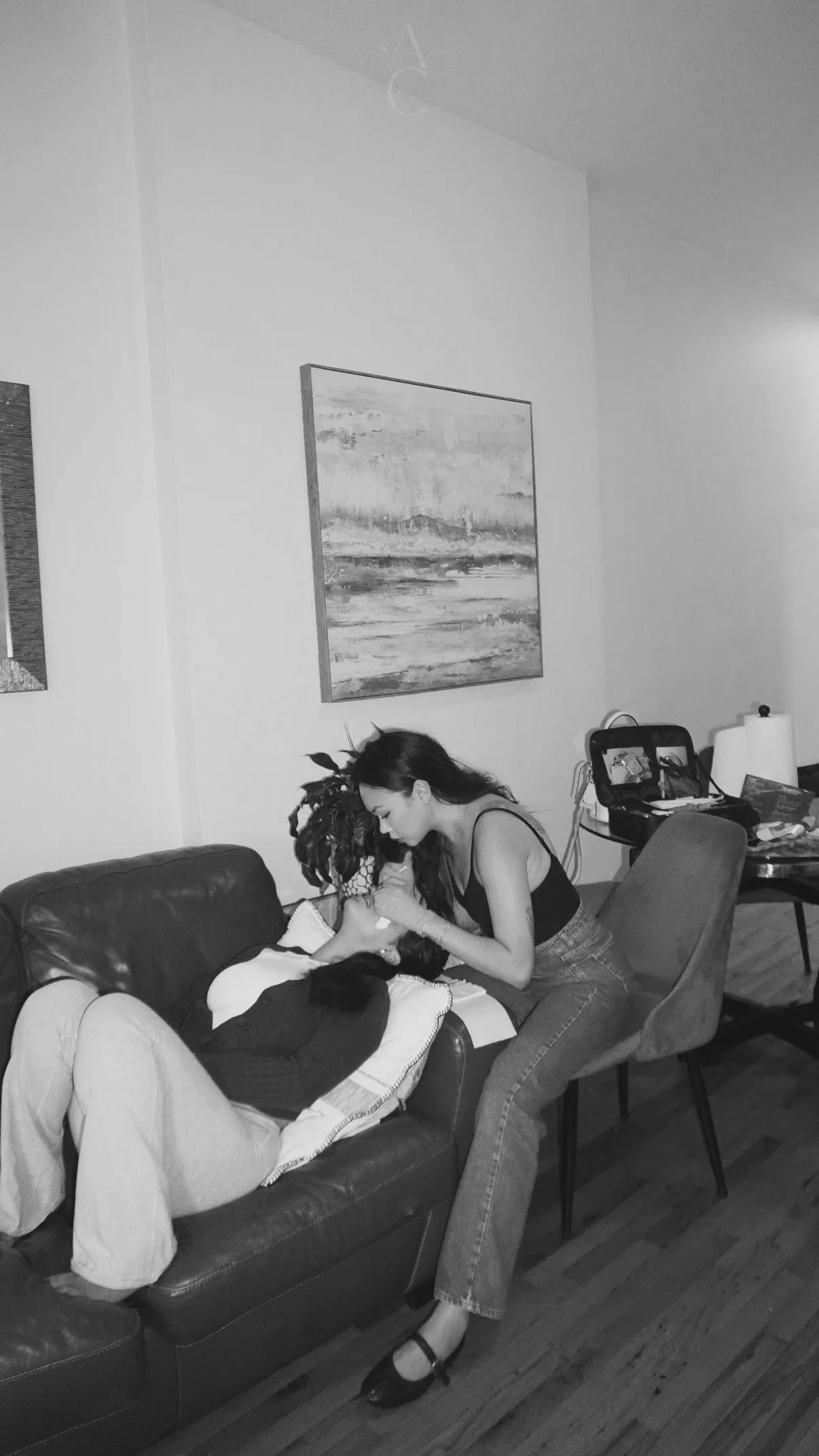 A woman leaning over a person lying on a couch, appearing to be breastfeeding in a living room with wall art, a table, and various items.
