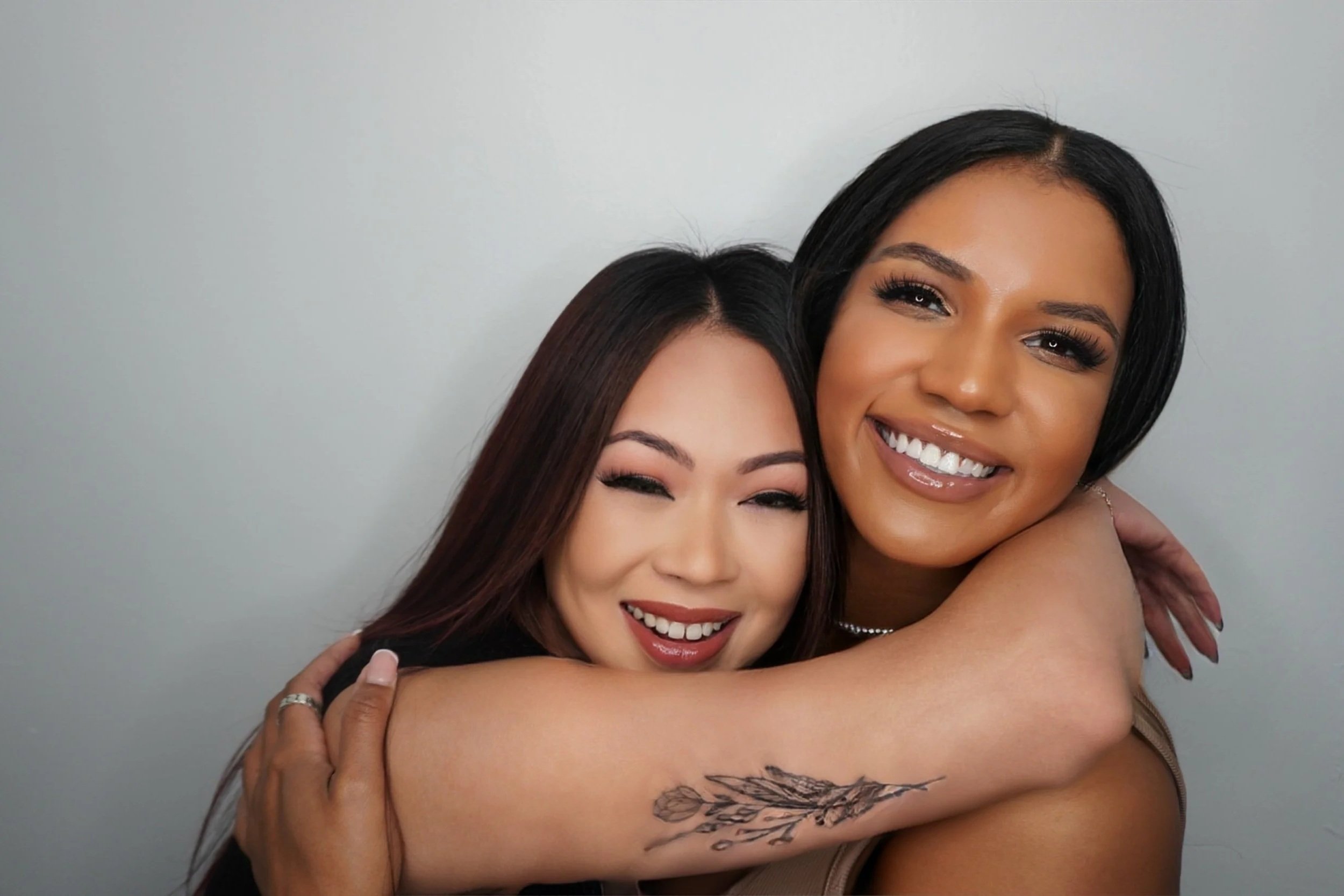 Two women smiling and hugging against a plain gray wall.