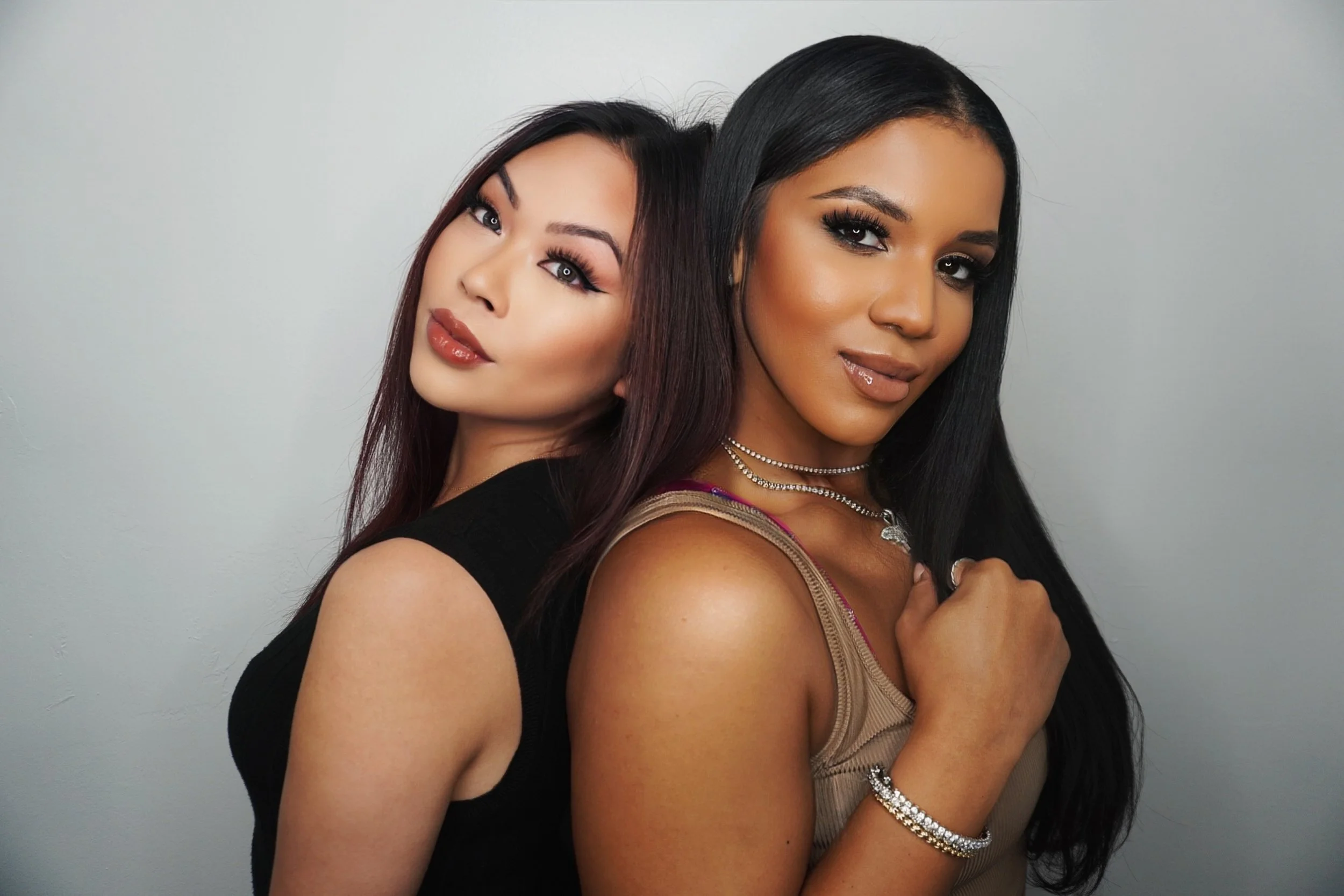 Two women posing closely together, one with long dark hair and the other with medium-length black hair, both wearing makeup and jewelry, against a plain light gray background.