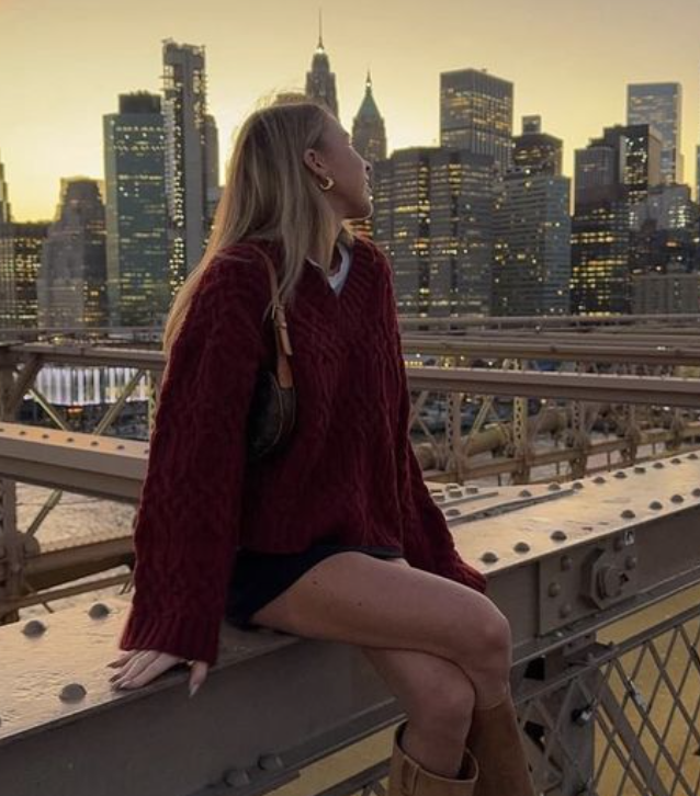 A woman sitting on a bridge with a city skyline at sunset in the background.