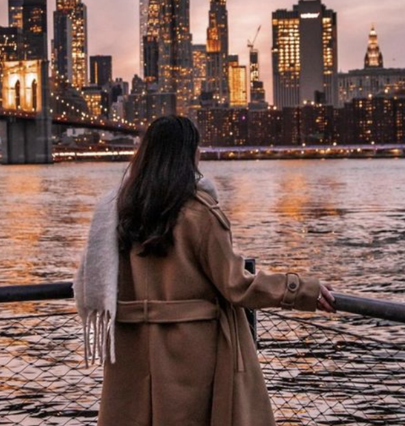Woman with long dark hair in a brown coat and scarf, standing by the water at sunset, overlooking the New York City skyline with tall lit buildings.