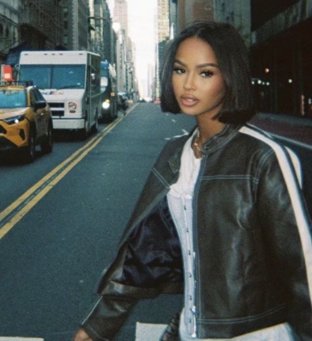 Young woman with short black hair and a leather jacket standing on a city street with yellow taxis and buses in the background.