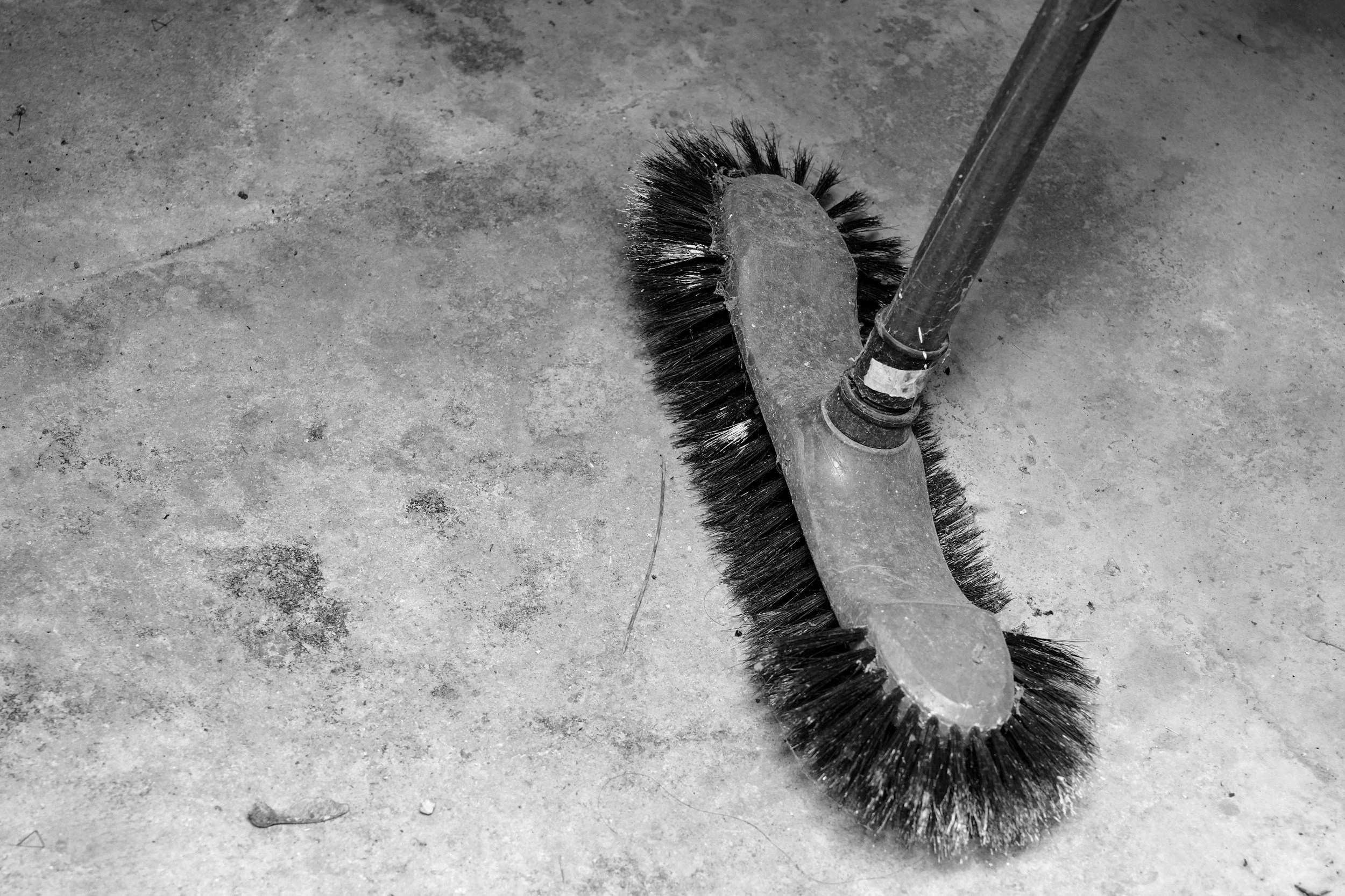 A floor cleaning brush with black bristles attached to a yellow handle, lying on a rough concrete surface.