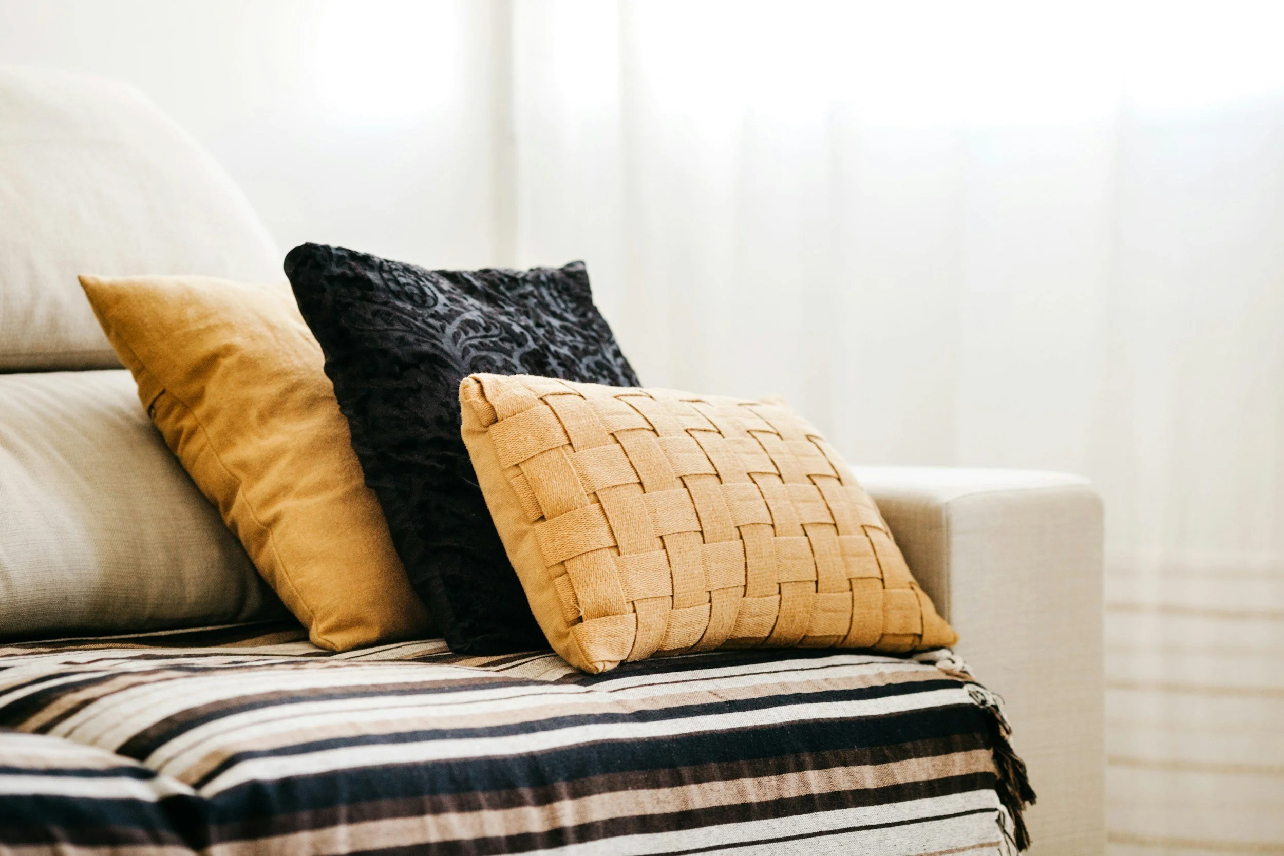 A collection of decorative pillows on a bed, including one black velvet, one mustard, and one woven beige pillow, with a striped blanket at the foot of the bed.