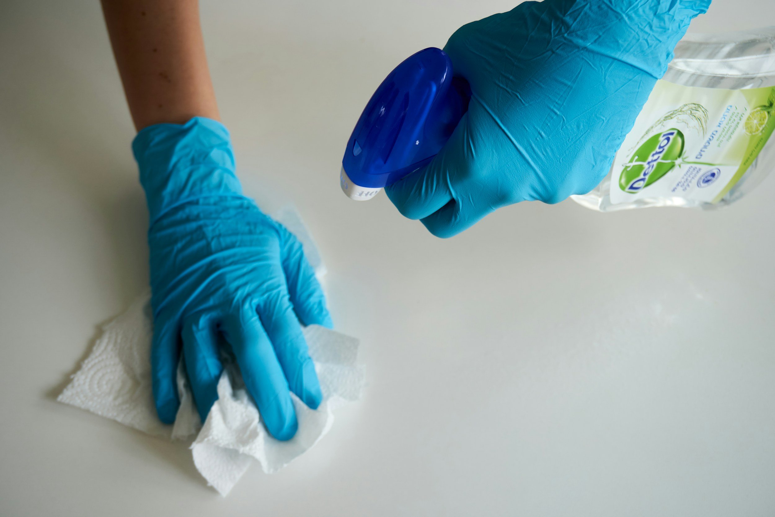 Person wearing blue gloves using a paper towel to wipe a surface. Hand sanitizer and a bag of disinfecting wipes are on the surface.