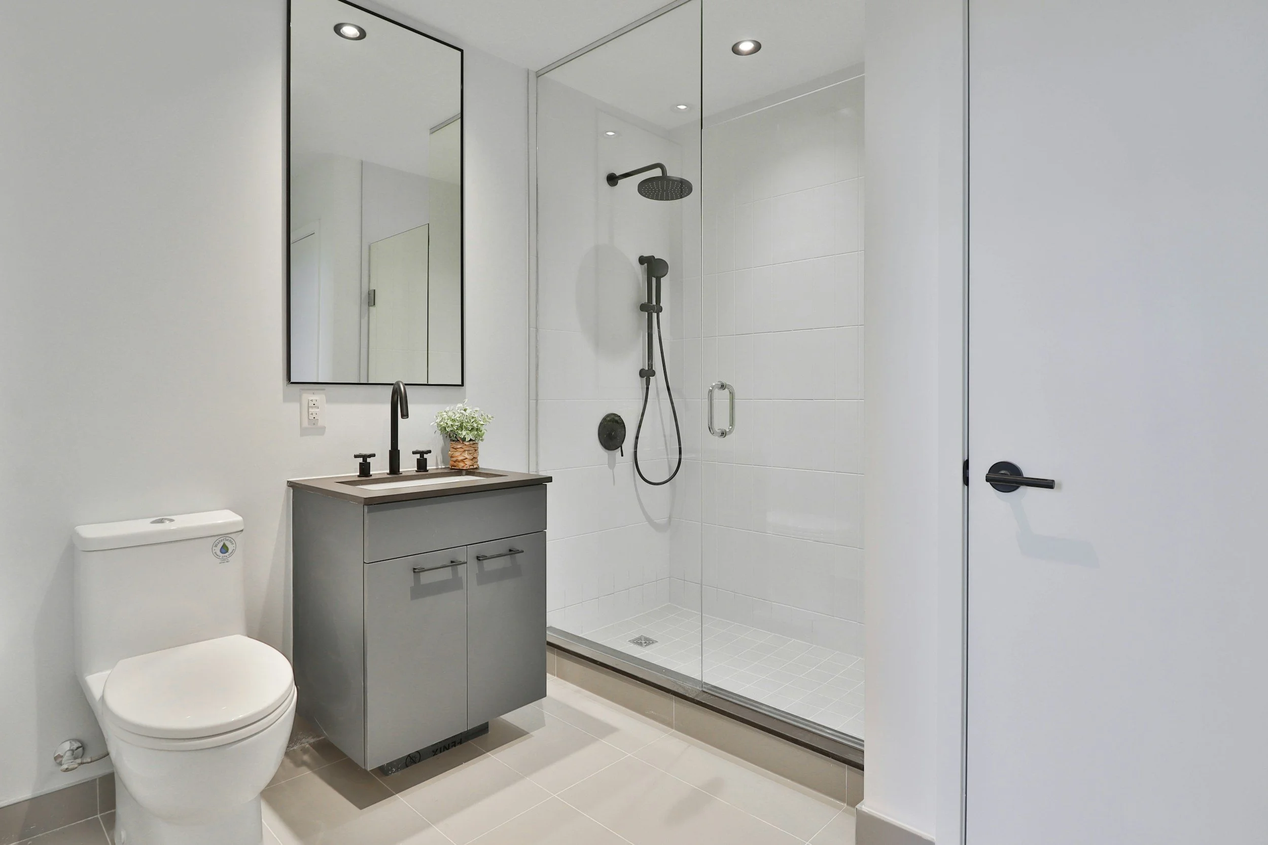 A modern bathroom featuring a white toilet, a gray vanity with black faucet, a potted plant, large mirror, and a walk-in shower with glass enclosure, white tiled walls, and black fixtures.