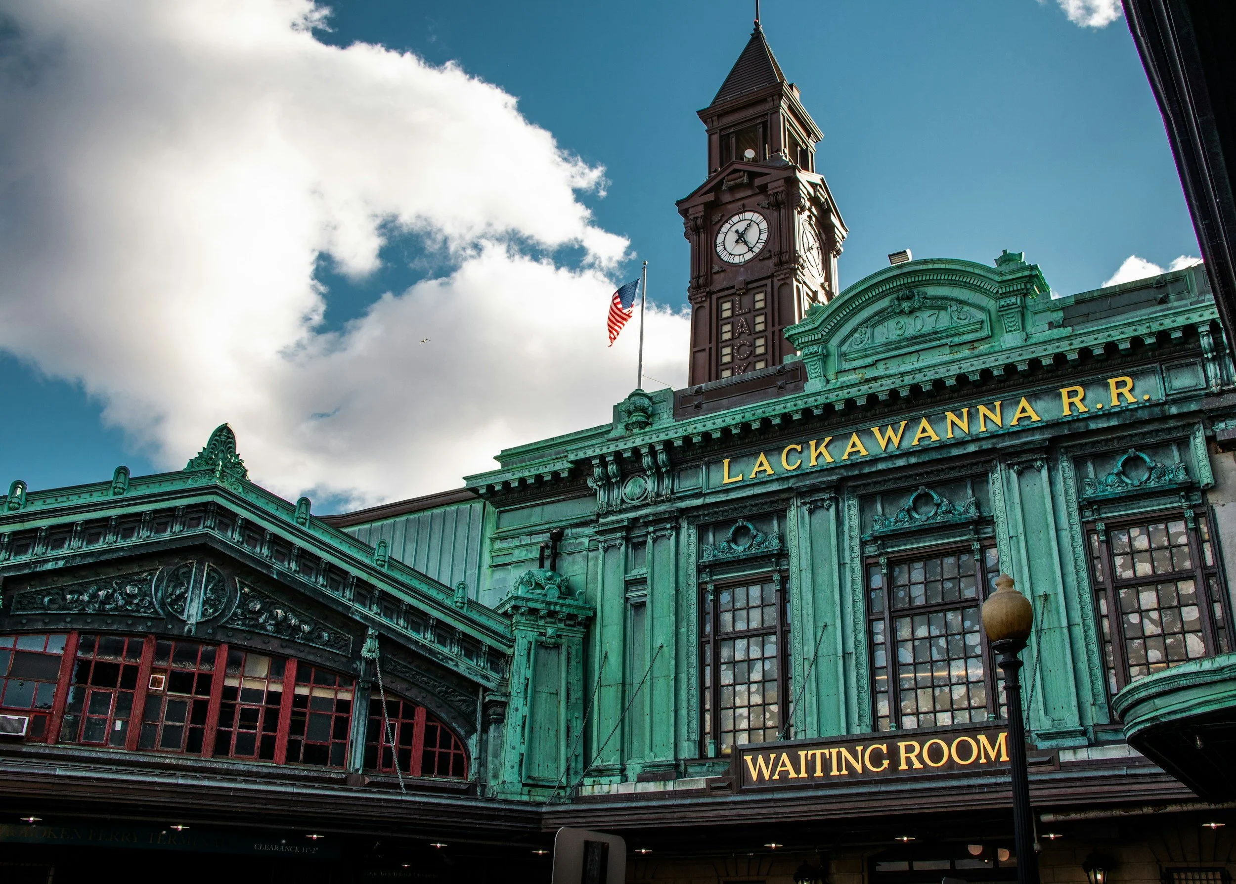 Historic building with a clock tower and the sign 'LACKAWANNA R.R.' on a green facade, featuring a waiting room and an American flag, under a partly cloudy sky.