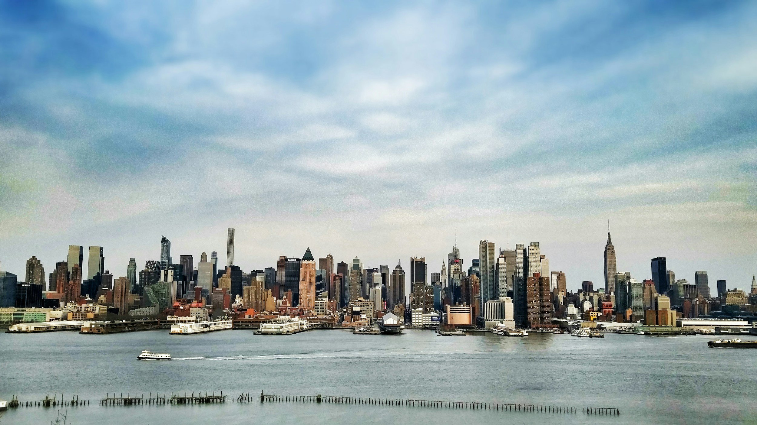View of a city skyline with numerous tall buildings, a waterway with boats, and a partly cloudy sky.