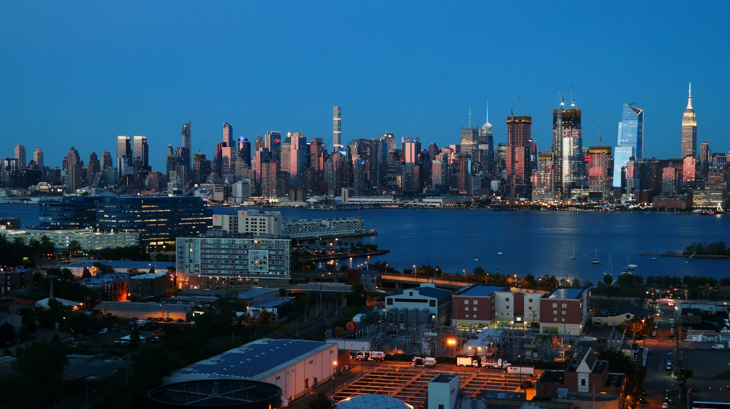 Nighttime view of New York City skyline with illuminated skyscrapers and water in the foreground.
