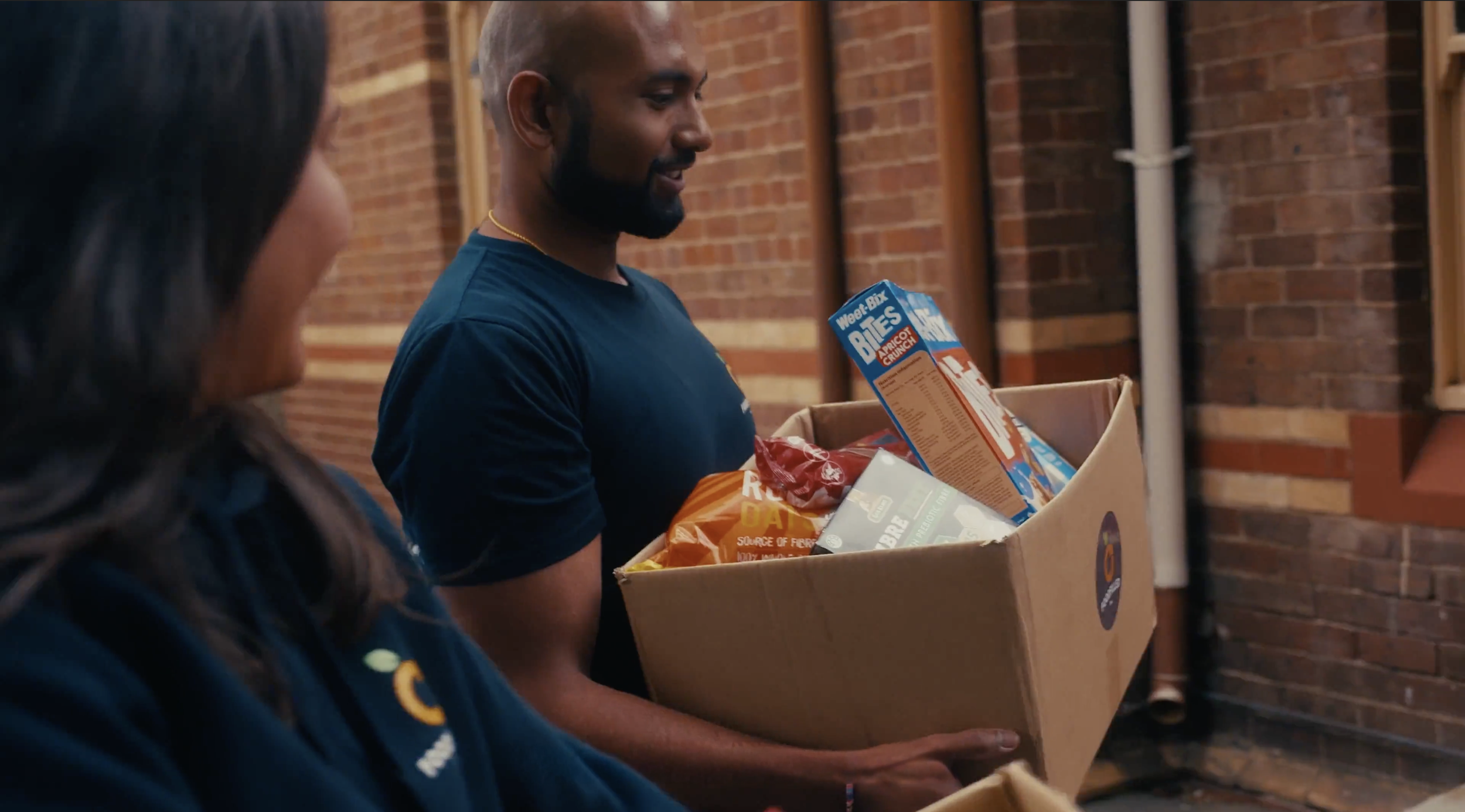 A man carrying a box of groceries, including cereal and snack items, standing next to a woman in an outdoor brick alleyway.