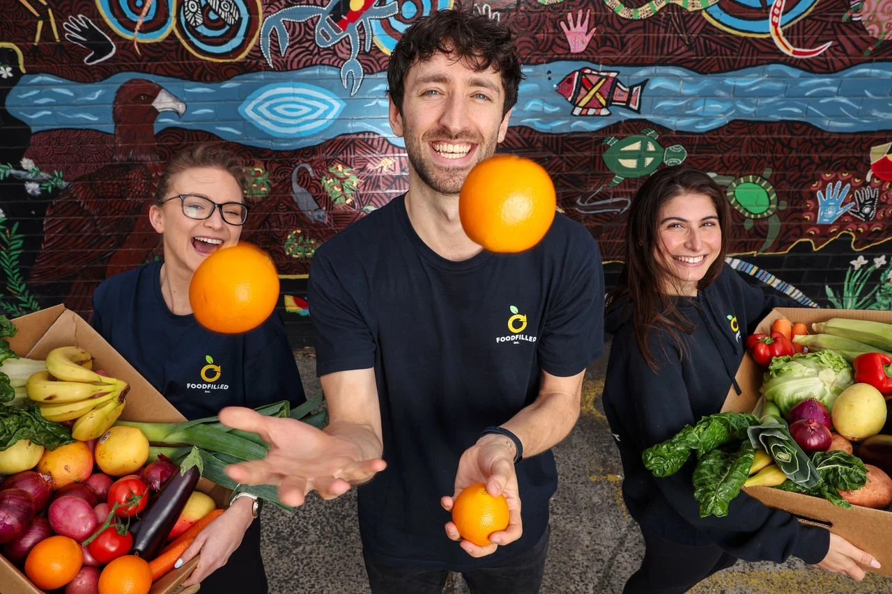 Three people wearing matching shirts, two women and one man, smiling and holding oranges and vegetables at a produce market with a colorful mural in the background.