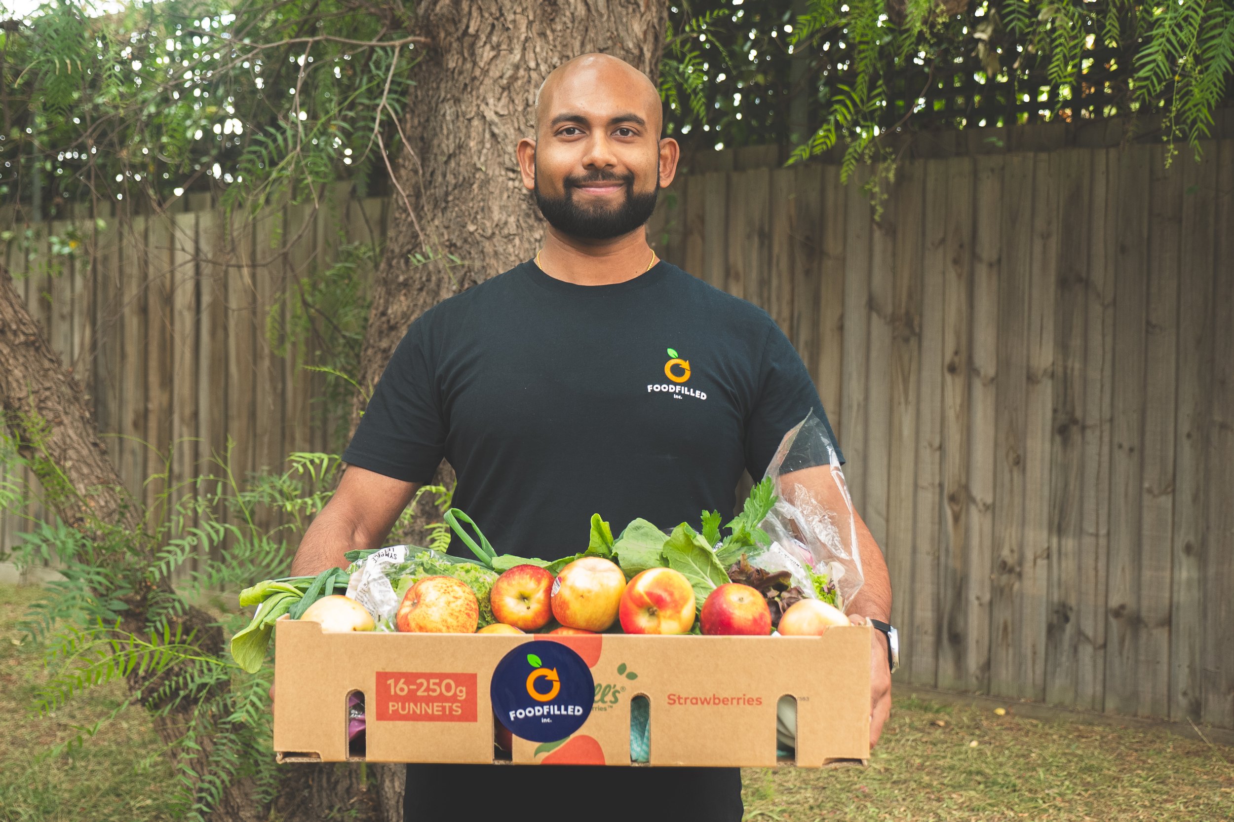 A man with a beard and bald head, wearing a black T-shirt with a Foodfilled logo, stands outdoors holding a box of fresh apples, strawberries, and greens. He is smiling and standing in front of a tree and a wooden fence.