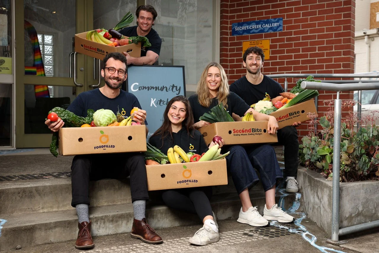 Group of five young adults smiling and holding cardboard boxes filled with fresh vegetables outside a community kitchen.