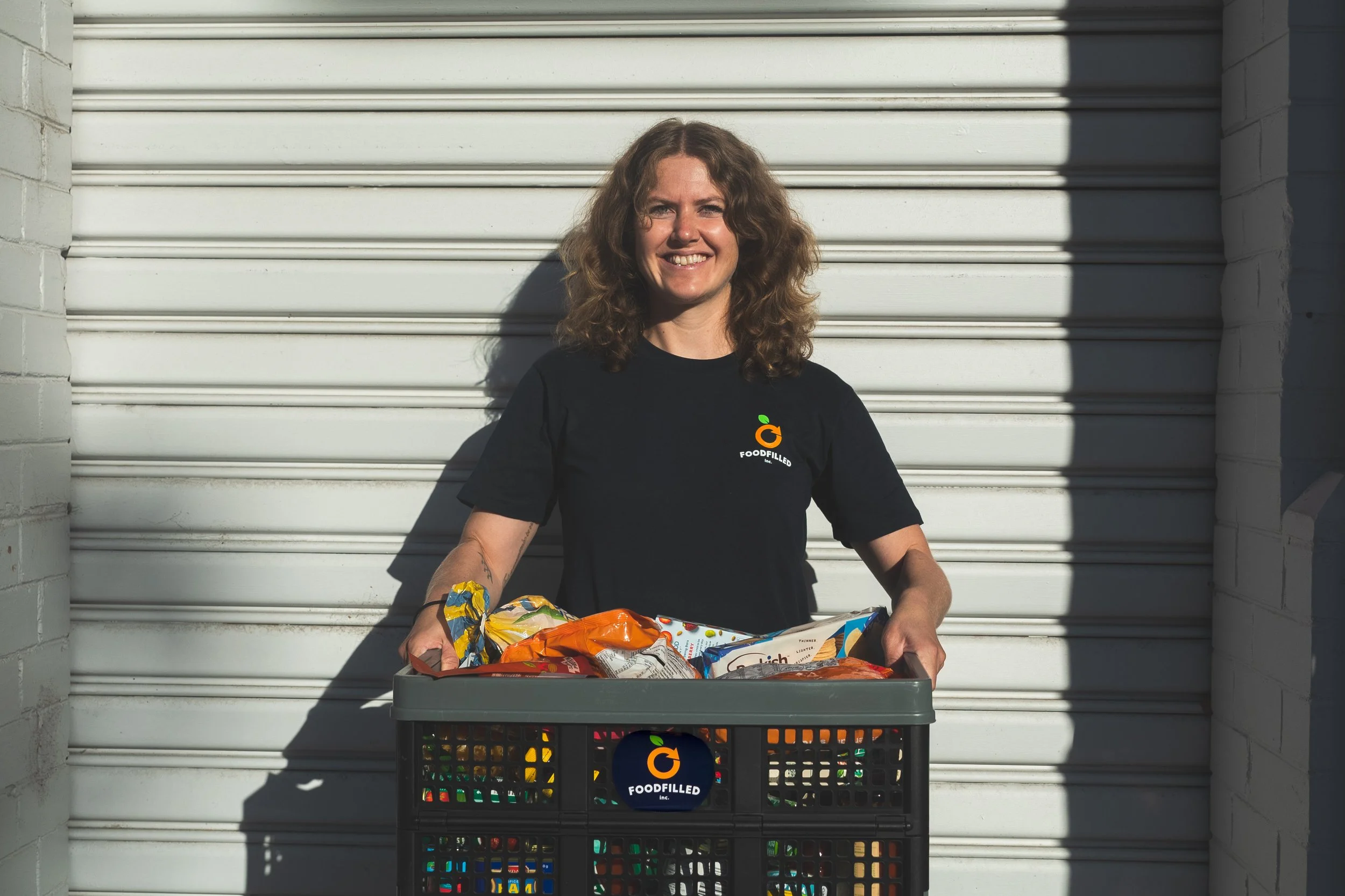 Woman smiling holding a crate filled with groceries in front of a white garage door