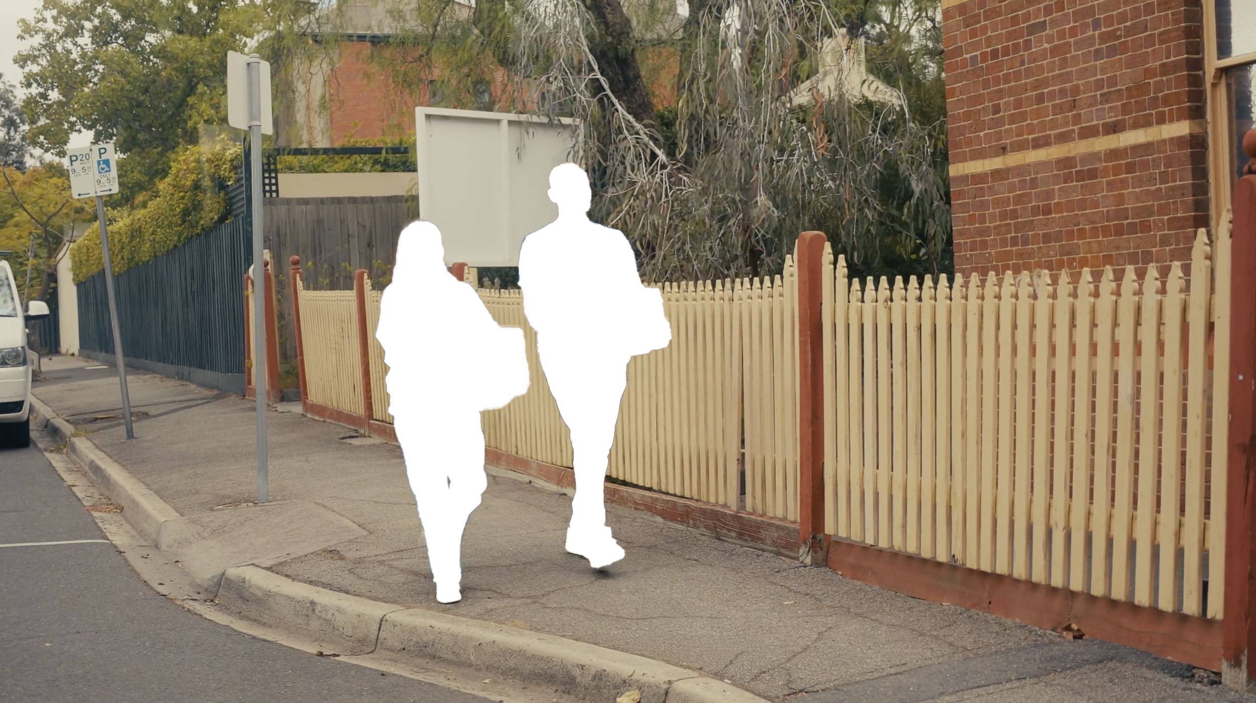 Two people walking on the sidewalk next to a wooden fence and brick building, with a street parking sign and a white vehicle parked nearby.