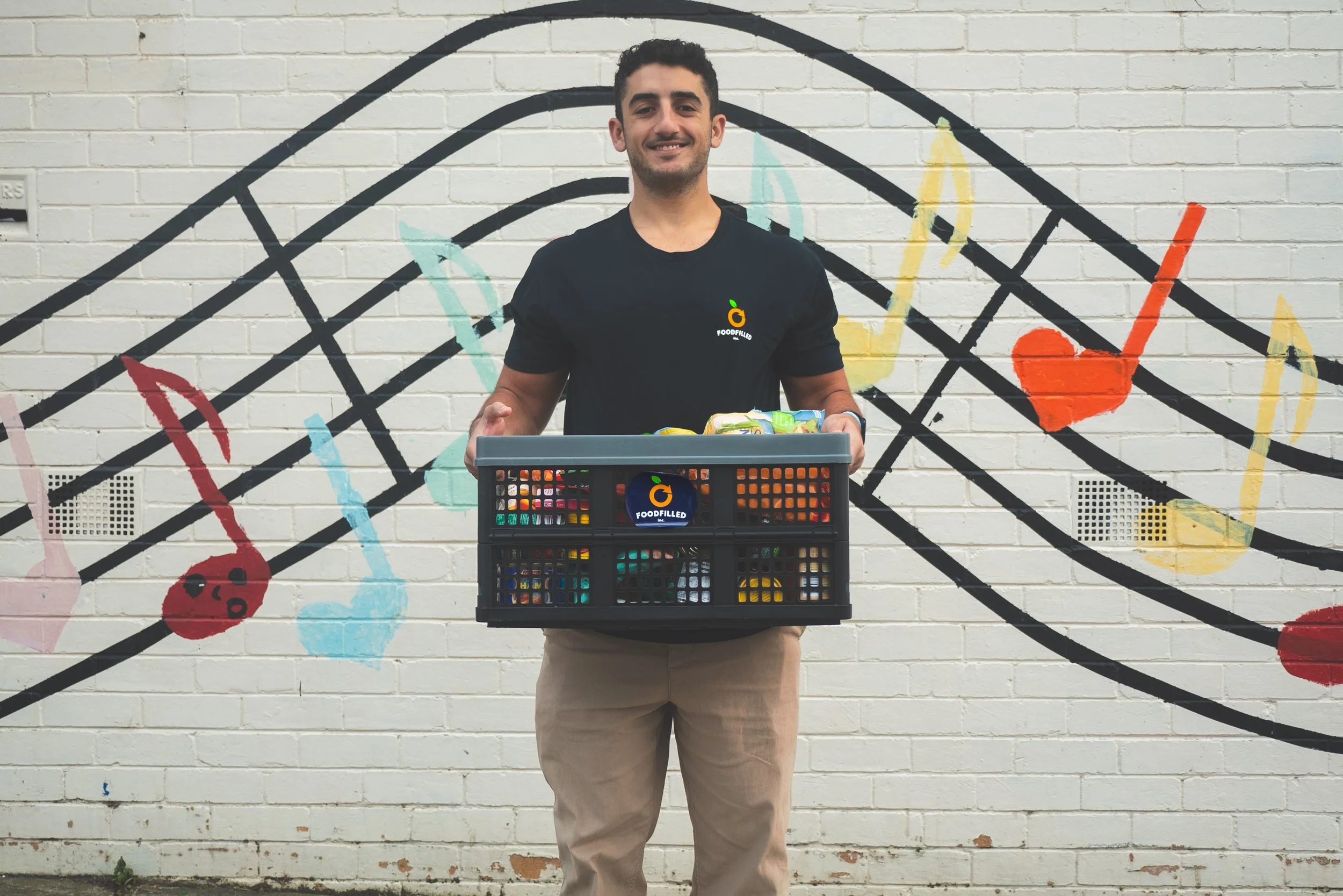 A man standing in front of a painted wall with musical note and heart designs, holding a black crate filled with groceries, wearing a black T-shirt with a food-related logo.