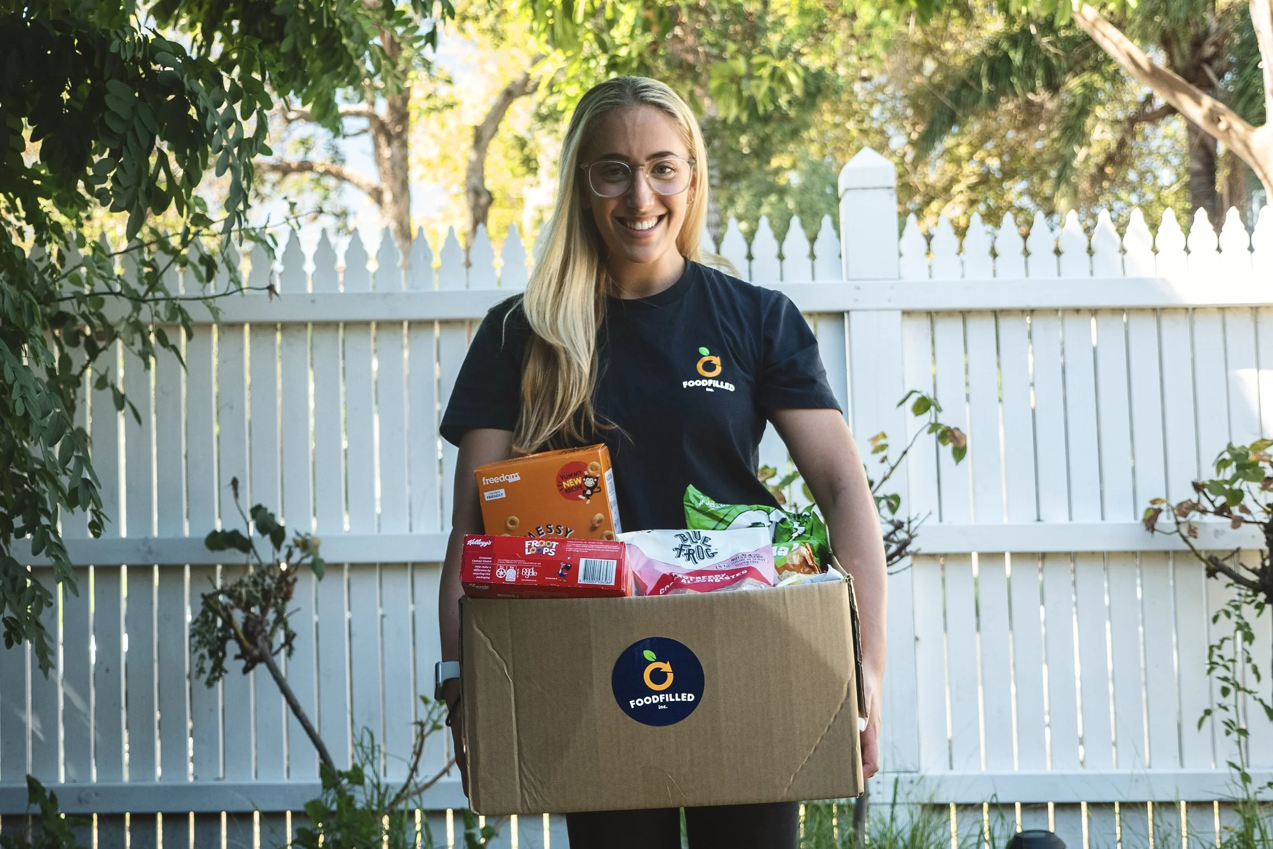 A smiling woman holding a cardboard box filled with groceries outdoors against a white picket fence and green trees.